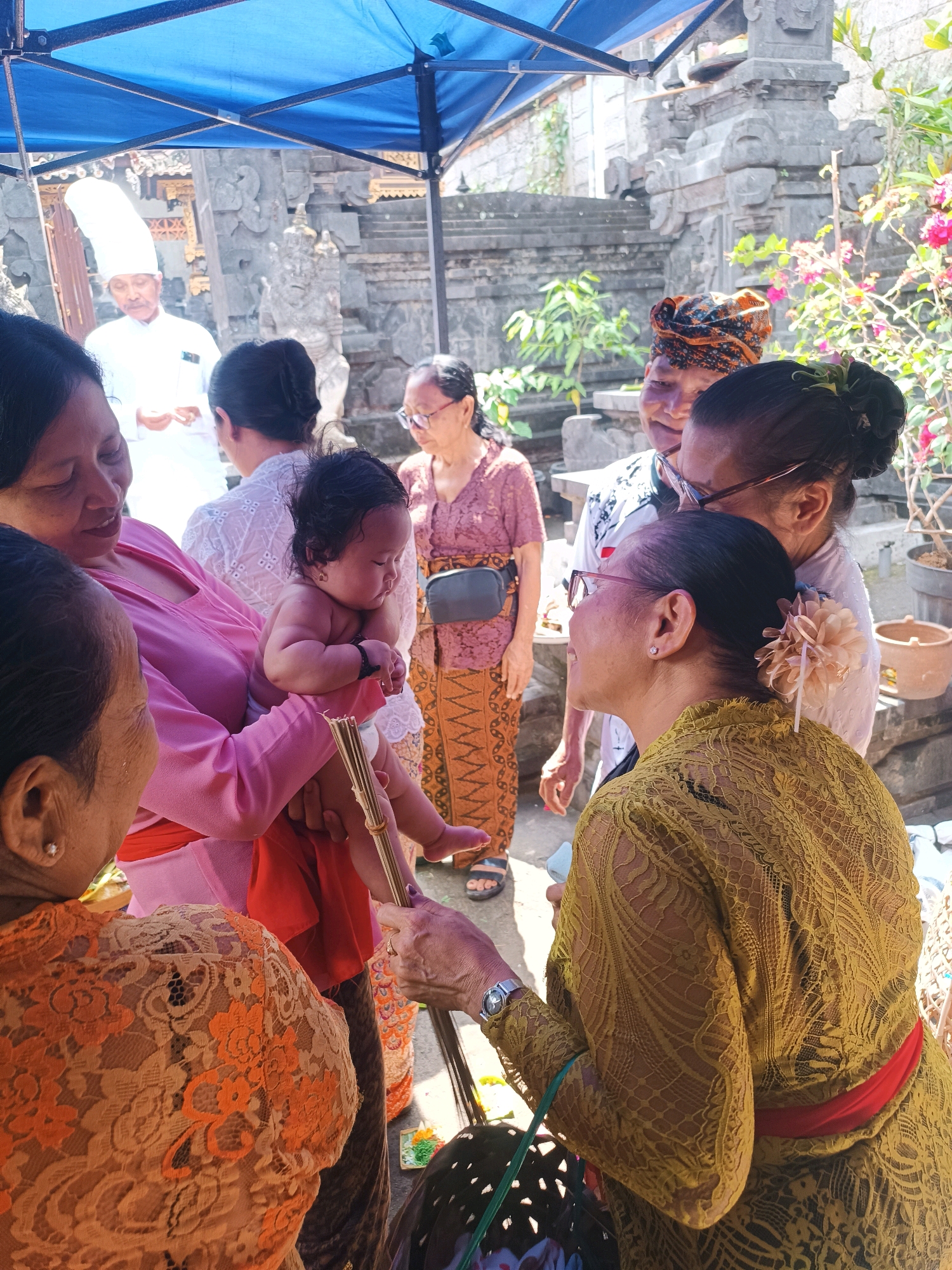 The image depicts a traditional Balinese ceremony taking place outdoors in the city of Tabanan, Indonesia. In the foreground, a woman in a vibrant yellow lace dress, cinched with a red sash, is holding a bundle of incense sticks. She is looking towards a baby being held by another woman in a pink blouse. The baby is naked and appears to be the focus of attention.
In the background, a man in a tall white headdress and white attire stands under a blue canopy, holding something in his hands, possibly a religious text or offering. Several other women, dressed in traditional Balinese clothing, are present, observing the proceedings. The environment features stone structures with intricate carvings, indicative of Balinese temple architecture, and lush greenery. The overall scene suggests a religious ritual or a cultural event, possibly a blessing ceremony for the infant. The sunlight casts strong shadows, indicating a bright, daytime setting.