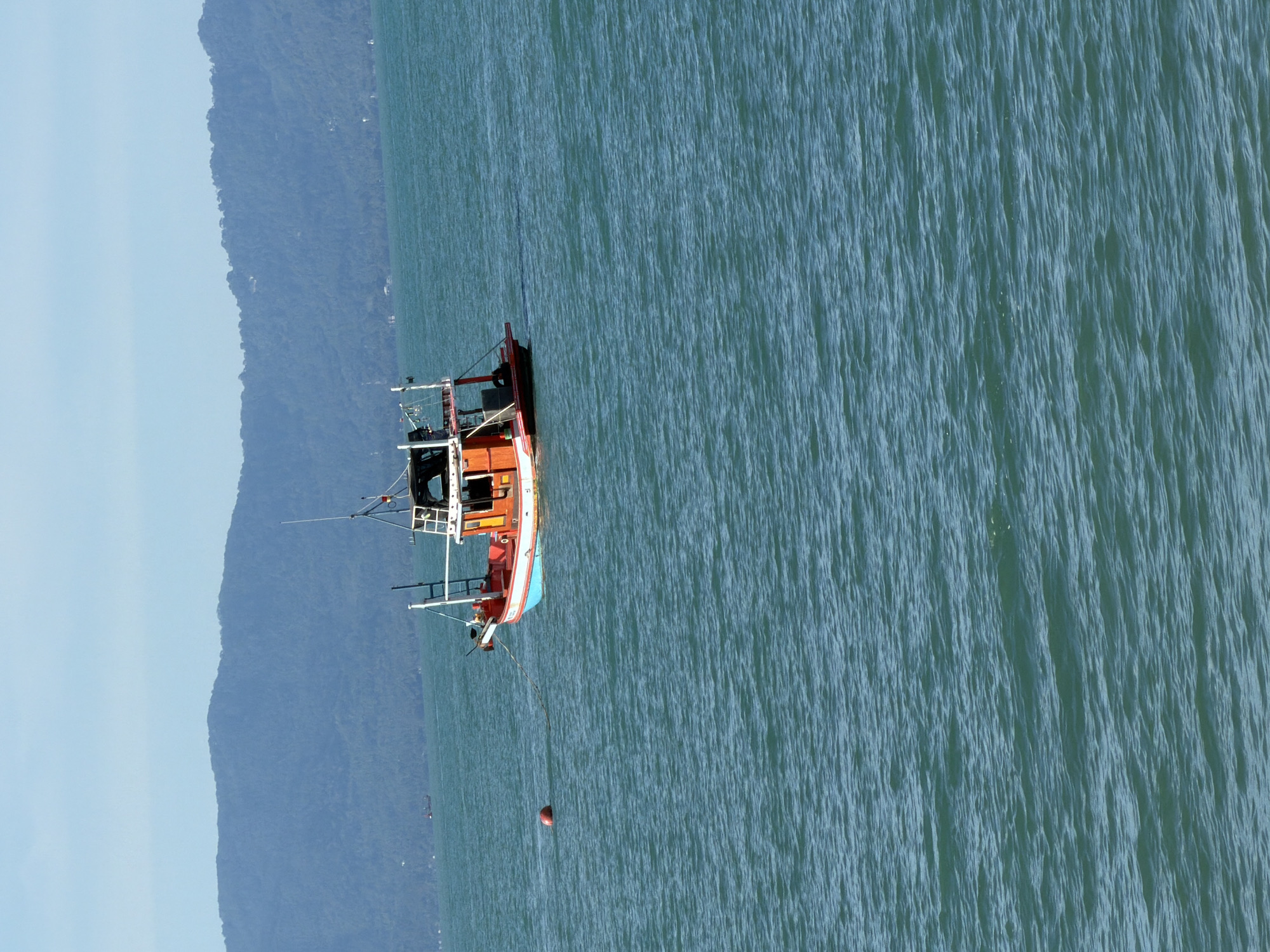 The image captures a fishing boat on the turquoise waters of Baan Tai, Thailand. The boat, painted in vibrant red and blue, is anchored with a red buoy. In the background, a lush green mountain range rises against a clear blue sky. The water is calm, with gentle ripples reflecting the sunlight. The scene is serene and tranquil, suggesting a peaceful day at sea.