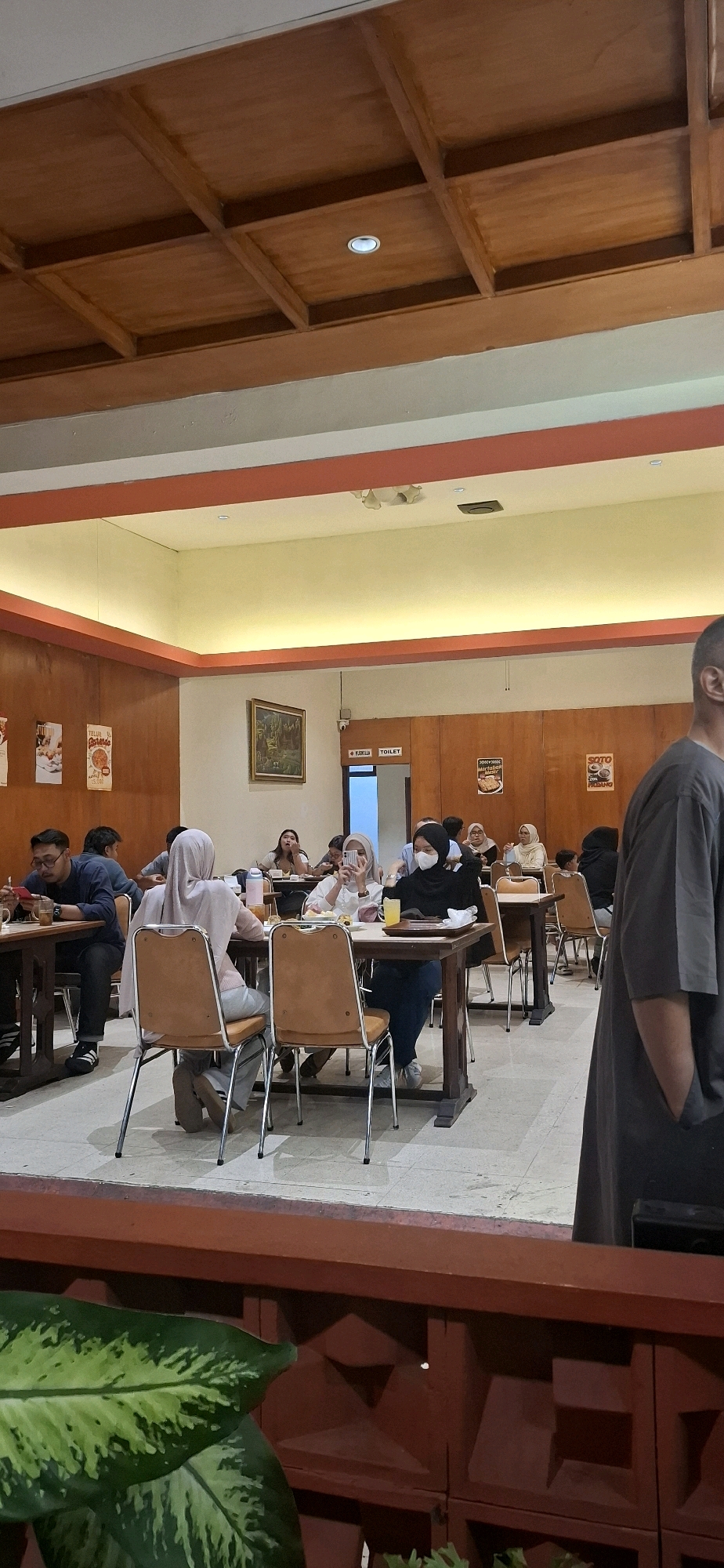 The image shows the interior of a restaurant with wooden paneling and a coffered wood ceiling with recessed lighting. Several tables are set up with chairs, and people are seated at some of them, eating and socializing. In the background, a sign indicates "TOILET." The overall atmosphere appears casual and communal. This scene is consistent with the dining environment found in many restaurants in Surakarta, Indonesia, known for its traditional Javanese cuisine and vibrant culinary scene.