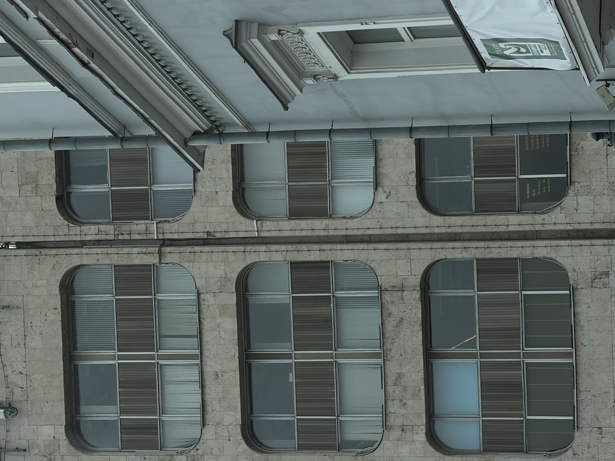 The image displays a detailed view of two adjacent building facades in Moscow, Russia, specifically the Moscow Art Theatre (MXAT) building at Tverskoy Boulevard 25.
The left building features a light-colored, textured stone-clad facade. Three large, horizontally aligned, rounded-rectangular windows are vertically stacked. Each window is divided into multiple panes by a grid pattern. The upper sections of the top two large windows have arched glass panes. Some window sections are fitted with vertical blinds, while others have white curtains. Darker, vertical paneling is present in the central, non-glass portions of these windows.
The right building, partially visible, has a smooth, light blue-grey painted facade. A grey metal downspout runs vertically along its left edge, separating it from the adjacent building. This facade includes a cornice adorned with a row of small, spherical light bulbs. Below the cornice, a rectangular window frame with ornate white architectural trim, featuring scrollwork and decorative patterns, is visible. Beneath this window or a balcony, a white banner with green text that reads "ТЕАТР" (Theater) and a stylized green "M" logo is displayed. No people are visible in the scene.