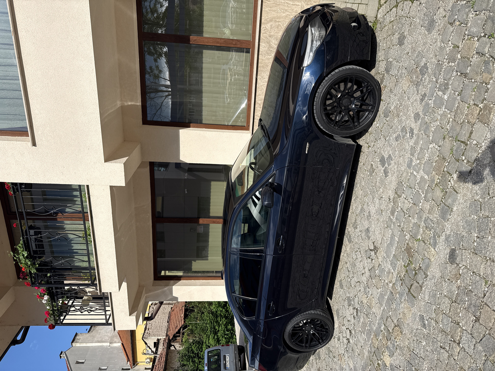 A black sedan is parked on a cobblestone street next to a cream-colored building. The building has dark-framed windows and a wrought-iron balcony with flowers. A partial view of adjacent buildings is visible in the background, including one with a red-tiled roof. In the far background, a utility vehicle is parked. The image was taken in Gabrovo, Bulgaria.