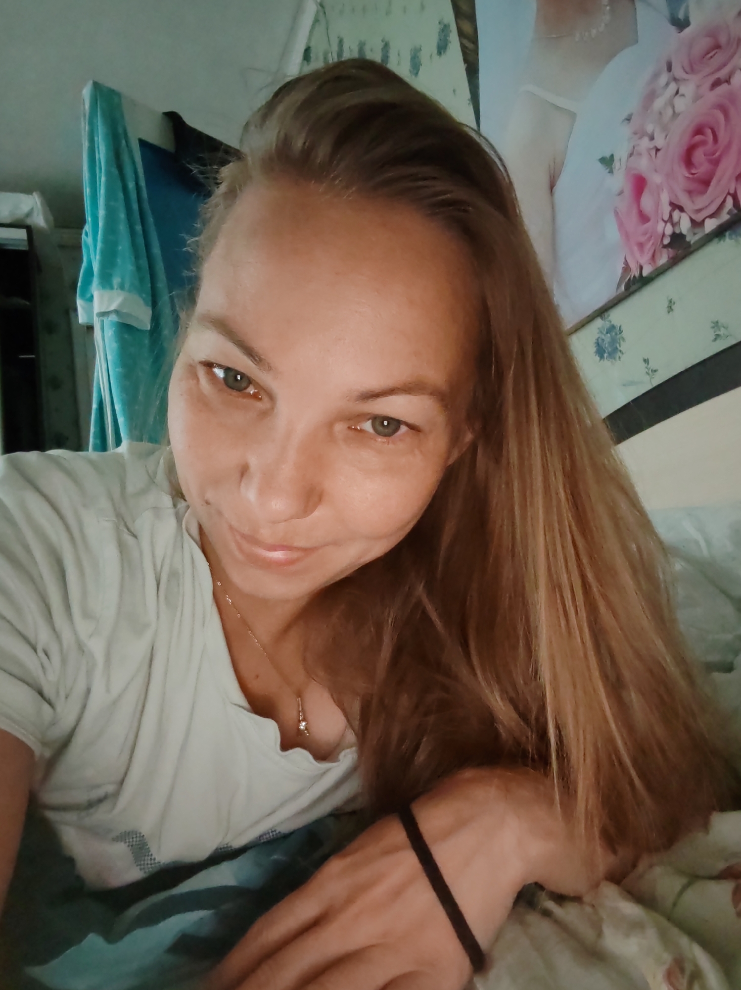 A woman with long, light brown hair is lying down, looking towards the camera. She is wearing a light-colored t-shirt and a necklace with a pendant. Her left arm is resting on the bed, and she has a dark hair tie on her wrist. Behind her, there is a blue towel hanging and a framed picture of a bride with a bouquet of pink roses. The overall setting appears to be a bedroom, likely in the city of Chuchlem, Russia.