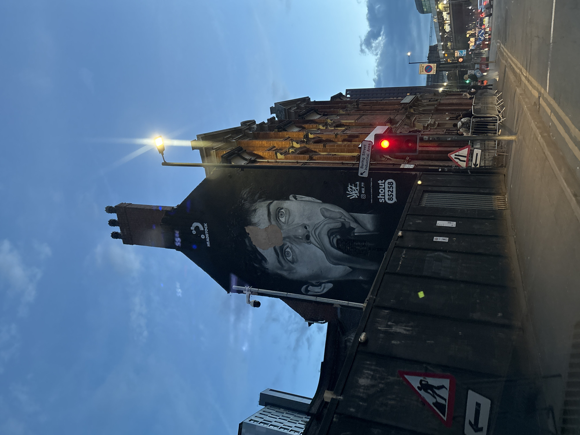 The image shows a street scene in Manchester, United Kingdom. A tall building with a large black and white mural of a distressed man's face dominates the left side of the frame. The mural includes the text "HEADSTOCK" and "shout 85258." A street lamp shines brightly next to the building.
On the right side, there is a street with a red traffic light, a "Baring Street Industrial Area" sign, and a "Men At Work" sign on what appears to be a large bin or container. A metal barrier is partially visible on the sidewalk. Several people are milling about, and some appear to be interacting. The sky is a mix of blue and dark clouds, suggesting it is evening or dusk. Other buildings and traffic lights are visible in the background.