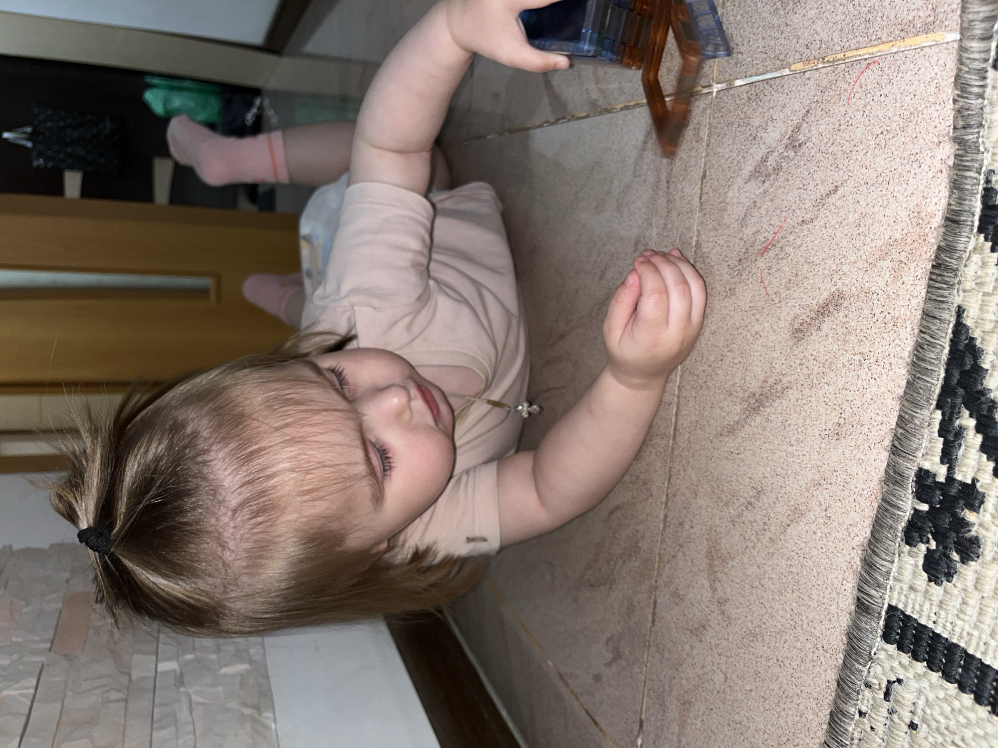 A young child lies on a tiled floor, playing with a transparent blue and orange toy. The child, wearing a pale pink t-shirt and pink socks, is on their side, looking towards the toy. Their blond hair is tied back with a black hair tie. A patterned rug is visible on the right side of the image, and a wooden dresser or cabinet is in the background. The scene appears to be indoors, possibly a home. The lighting suggests it might be evening or the room is dimly lit. The child's expression is one of concentration and engagement with their toy. There is no visible text in the image. The location is Novokuybyshevsk, Russia.