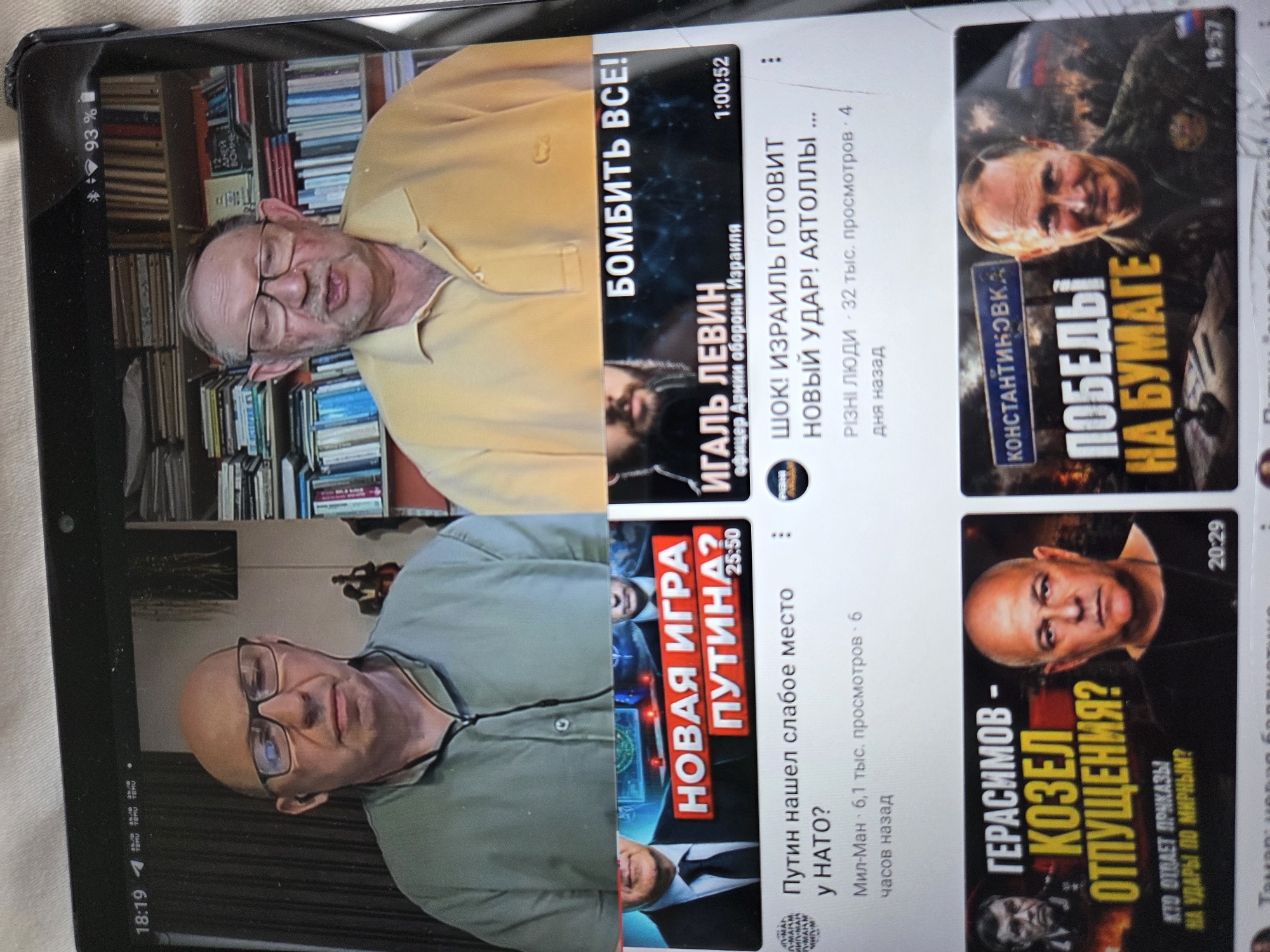 The image displays a tablet screen showing a YouTube interface with multiple video thumbnails. At the top, two men are visible, framed from the chest up, in what appears to be a video call or recorded segment. They are seated against backgrounds of bookshelves filled with books. Below these, three distinct video thumbnails are presented.
The first thumbnail is a news-style graphic with red text that reads "НОВАЯ ИГРА ПУТИНА?" (Putin's New Game?). Below this, a title reads "Путин нашел слабое место у НАТО?" (Did Putin find NATO's weak spot?).
The second thumbnail features a man with a beard and another figure obscured. The text on this thumbnail reads "БОМБИТЬ ВСЕ!" (Bomb Everything!) and "ИГАЛЬ ЛЕВИН" (Igal Levin) followed by "офицер Армии обороны Израиля" (Officer of the Israel Defense Forces). The video title below this thumbnail is "ШОК! ИЗРАИЛЬ ГОТОВИТ НОВЫЙ УДАР! АЯТОЛЛЫ ..." (Shock! Israel is preparing a new strike! Ayatollahs...).
The third thumbnail has a graphic with two men. The larger text reads "ГЕРАСИМОВ - КОЗЕЛ ОТПУЩЕНИЯ?" (Gerasimov - Scapegoat?). The text below asks "КТО ОТДАЕТ ПРИКАЗЫ НА УДАРЫ ПО МИРНЫМ?" (Who gives orders for strikes on civilians?).
The fourth thumbnail shows Vladimir Putin with text that reads "КОНСТАНТИНОВКА" and "ПОБЕДЫ НА БУМАГЕ" (Victories on Paper).
The context provided by the "Israel Defense Forces" mention in the second thumbnail, along with the presence of an Israeli officer's name, suggests that this content likely relates to geopolitical events involving Israel. However, the image itself does not provide any direct visual information about the city of Rishon LeZion, Israel.