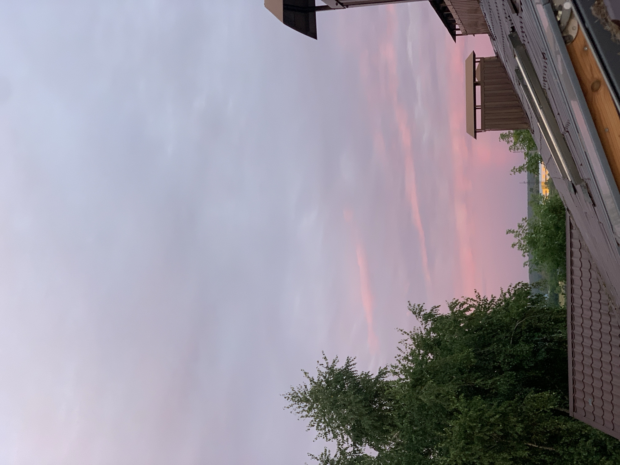 The image depicts a rooftop view in Hostomel, Ukraine, during what appears to be sunset. The sky is a soft gradient of pink and purple hues, with streaks of lighter pink clouds scattered across it. In the foreground, a brown-tiled roof with skylights is visible, showing signs of wear and tear. To the left, a lush green tree with dense foliage dominates the frame, its leaves catching the soft light. To the right, parts of two chimneys are visible, one made of dark wood and the other a darker structure with a metal cap. In the distance, faint outlines of trees and buildings can be seen against the horizon, with a bright yellow structure partially visible. The overall impression is one of a quiet, rural or suburban setting at the end of the day.