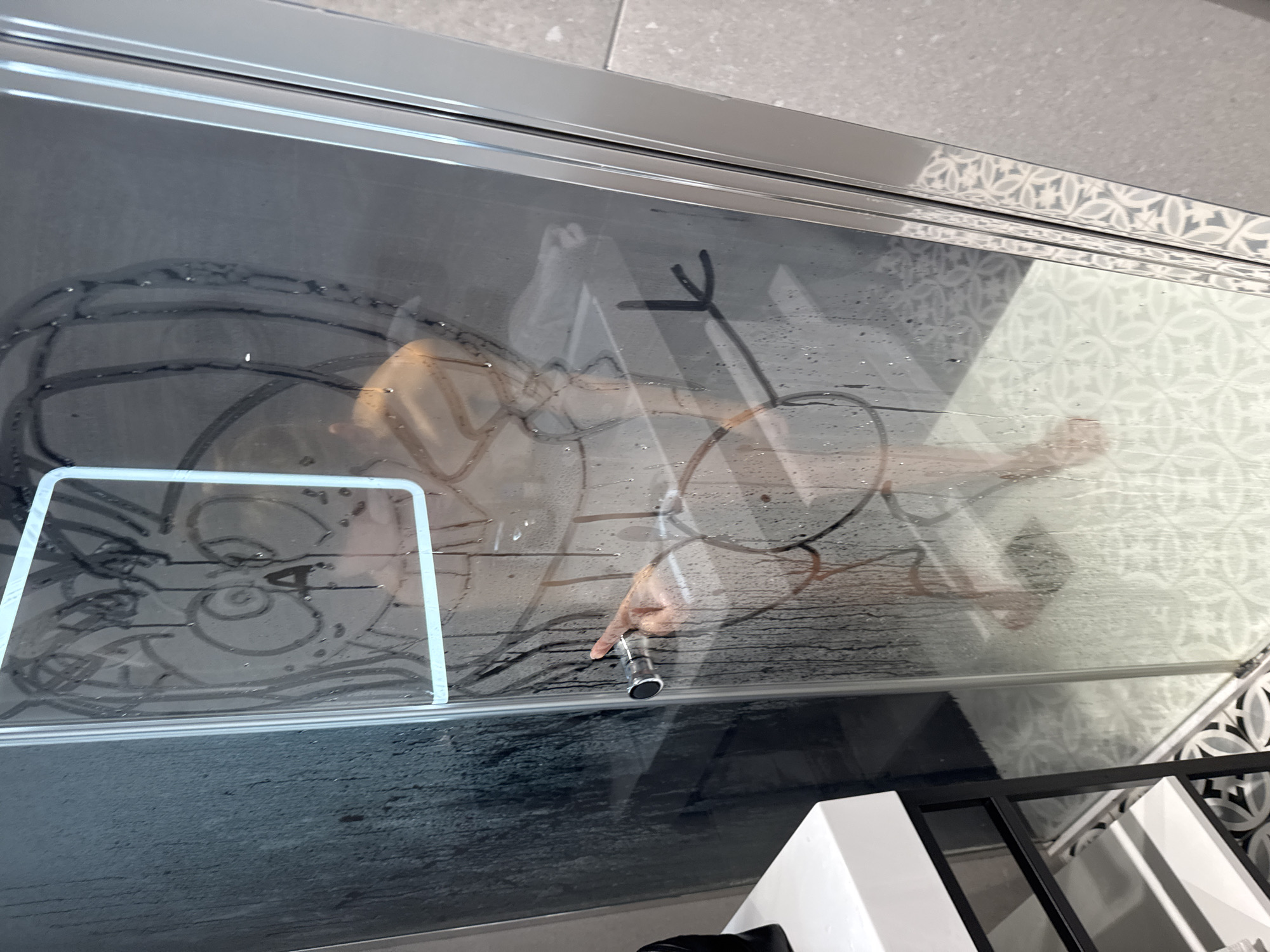 The image captures a view through a steamy glass partition, likely a shower door, into a bathroom. A person, appearing to be a child, is inside the enclosure, actively drawing on the condensed glass surface with their finger.
**Subjects:** The primary subject is a person, whose reflection or presence is visible behind the glass. Their face is partially obscured but discernible on the left side, and their hand is clearly visible on the right, holding a chrome fixture.
**Objects:** The central object is the large glass panel, covered in condensation and water droplets. Several drawings have been made on this surface: on the left, within a lighter rectangular outline, are circular shapes resembling eyes or a face; on the right, a stick-figure-like drawing (head, partial body, and extended arm) and a V-shaped mark are visible. The person's right hand is holding a polished chrome showerhead or similar water dispensing fixture. Behind the glass, on the left, a coiled black object resembling a hose or cable is present. The glass panel is framed by metallic rails. Outside the enclosure, on the right side, a decorative tiled wall with a grey and white geometric pattern is visible. In the bottom right corner, a white and black structure, possibly part of a vanity or shelving unit, can be seen.
**Scene/Setting:** The setting is clearly a bathroom, with the glass partition indicating a shower or bath enclosure. The presence of condensation suggests a humid environment, typical of a recently used shower.
**Activity or event taking place:** The person inside is engaging in a playful activity, drawing on the steamy glass. They are also holding a shower attachment, which might be in use or simply being held while they draw.
**Notable details:** The steam on the glass is thick enough to allow for drawing. The artistic style of the drawings is simple and childlike. The tiled wall outside the shower adds a decorative element to the bathroom. There is no visible text in the image. The lighting suggests an indoor setting, and the time of day is not discernible.