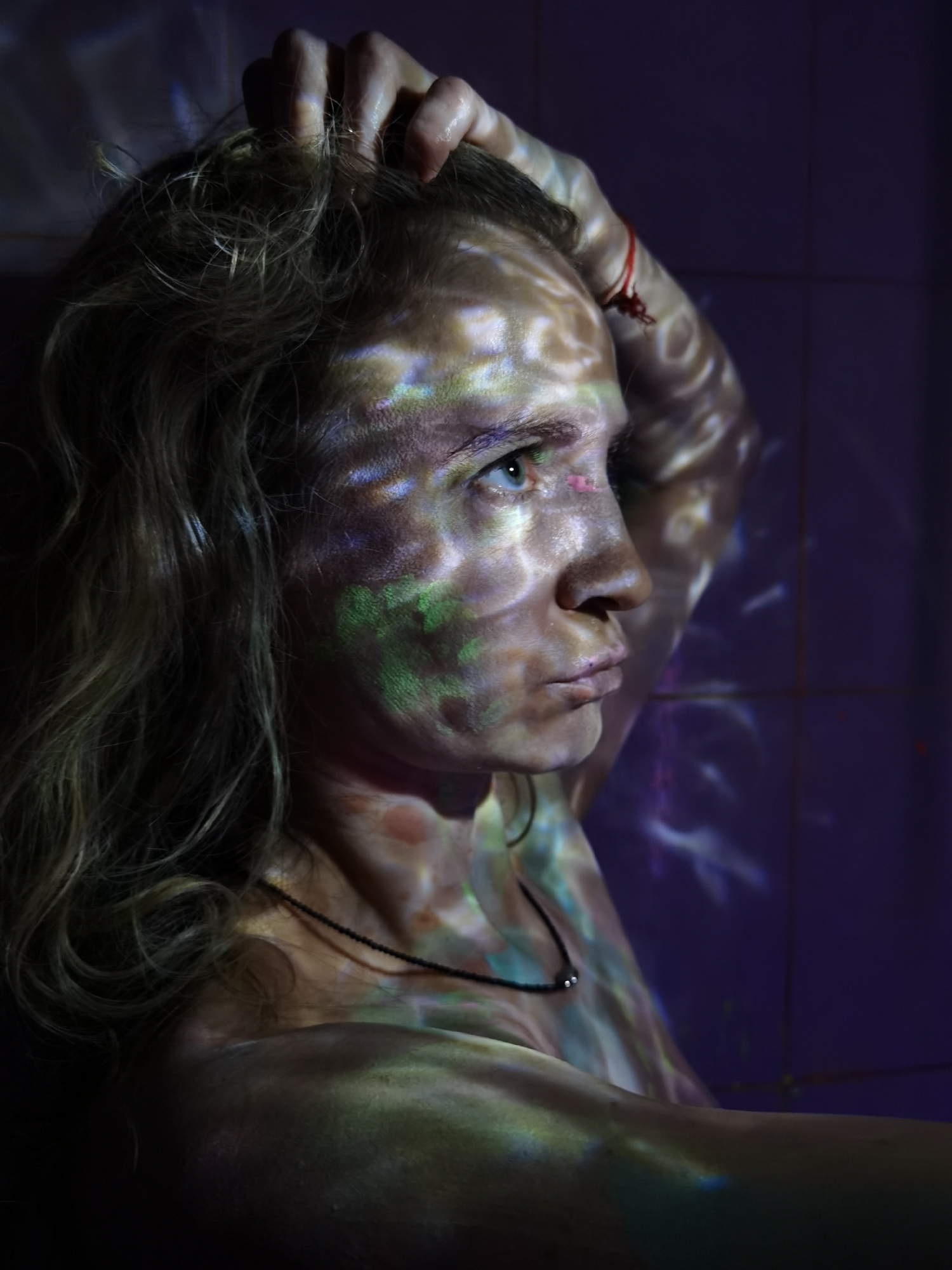 An artistic portrait captures a person, likely an adult female, in a profile view from the chest up, set against a dark, possibly tiled, background in shades of deep purple. Her light, wavy hair cascades around her shoulders, and one hand is raised, grasping her hair at the top of her head. Her face, illuminated by projected patterns of light, shows a contemplative or pensive expression, with her gaze directed forward and slightly upwards.
The most prominent feature is the dynamic interplay of light and color. Intricate, swirling patterns in various hues of green, blue, purple, yellow, and white are projected onto her skin, creating an ethereal and abstract visual effect across her face, neck, shoulder, and arm. Similar luminous patterns are visible on the dark background. A thin black necklace with a small, circular pendant adorns her neck. A small red string or bracelet is visible on her raised arm, near the wrist. The lighting suggests an indoor setting, likely dark, with the primary illumination originating from the projection. There is no visible text.
