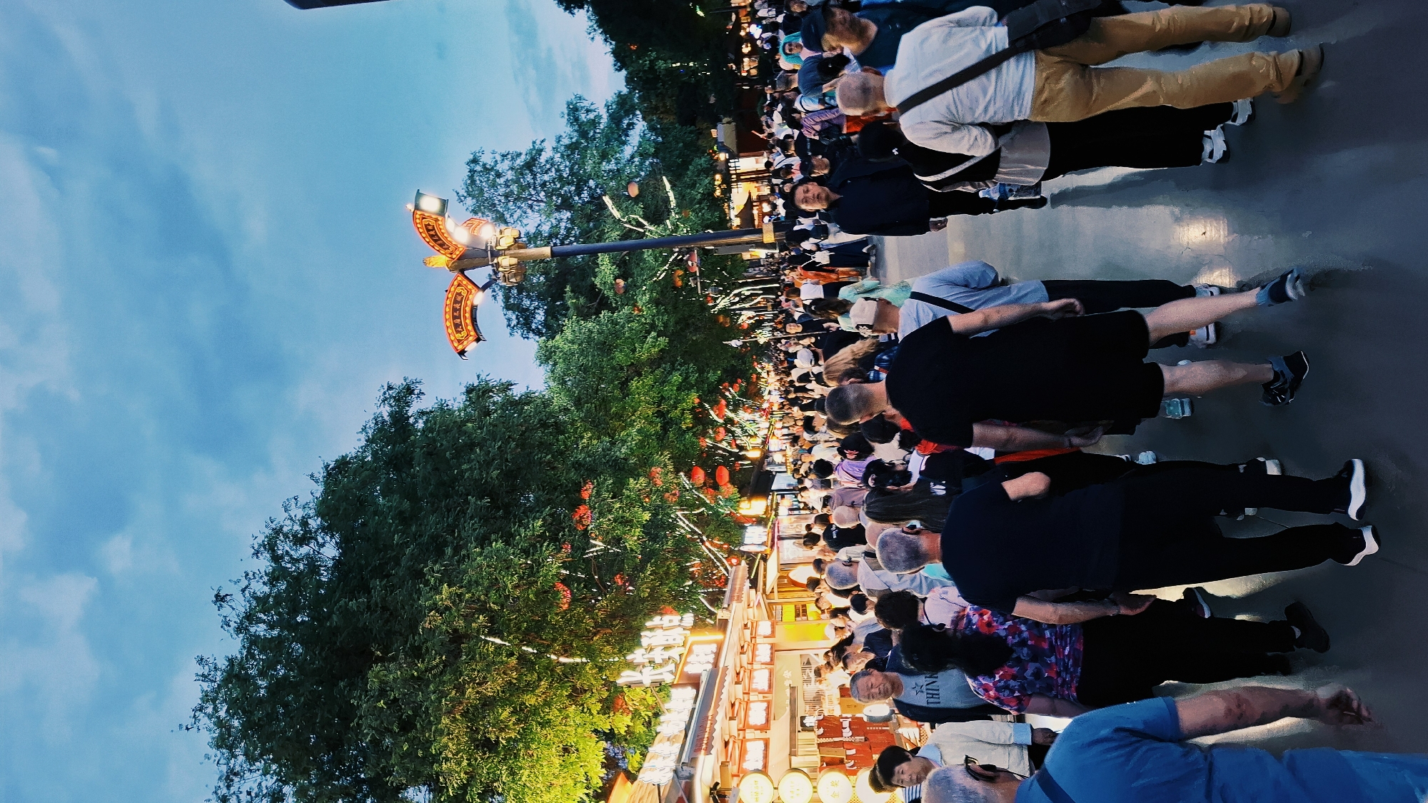 A street scene in Yanta District, China, shows a large crowd of people gathered. The scene features food stalls on the lower left, illuminated by lights. Above the stalls, trees and red lanterns are visible, leading to a streetlamp with decorative elements. The sky is visible above the scene, appearing blue with some cloud cover. The crowd of people extends into the distance, with the backs of their heads and shoulders being the primary visual focus. People are wearing various outfits.