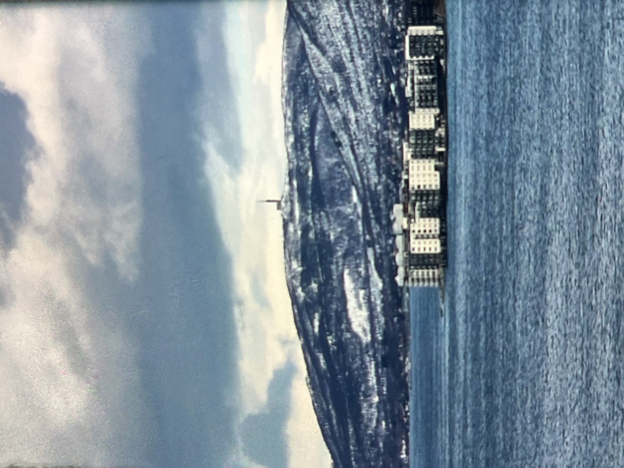 The image shows a body of water in the foreground, with a large, multi-story apartment building constructed on stilts or a pier extending into the water. The building has a modern design with many windows and balconies. In the background, a snow-covered mountain range rises, with a tall communication tower visible on its peak. The sky above is cloudy, with a mix of dark and light clouds. The overall impression is of a winter landscape in a coastal or lakeside city.