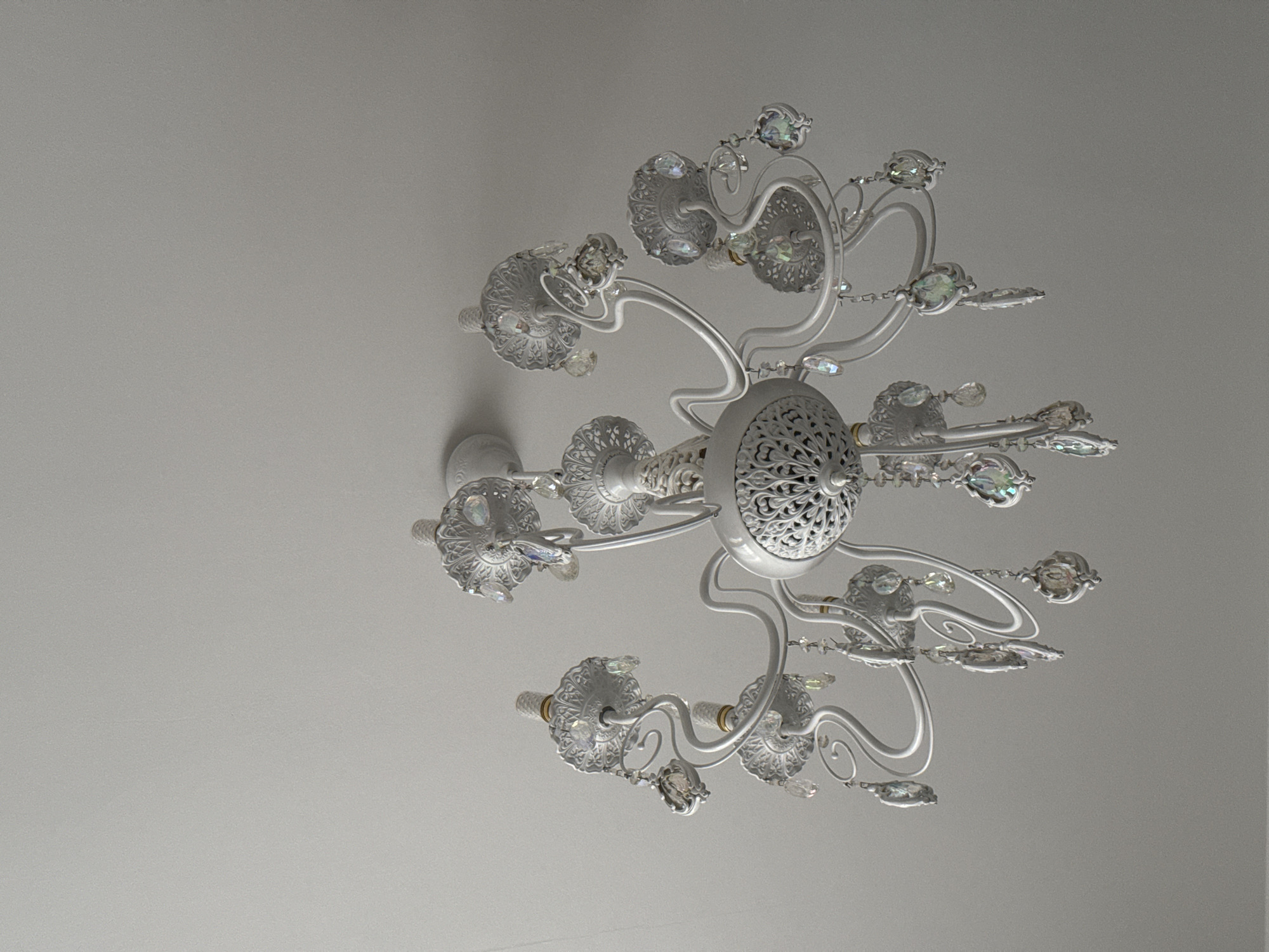 The image shows a white chandelier with multiple arms and ornate detailing. Crystal pendants hang from the arms, catching the light and creating a prismatic effect. The chandelier is suspended from a plain white ceiling. The lighting is soft and even, suggesting an indoor setting. The photograph appears to be taken from below, looking upwards at the chandelier. There are no people or other objects visible in the frame. The overall impression is one of elegance and a classic decorative style. The image does not provide any specific information to determine its location in Tashkent, Uzbekistan.