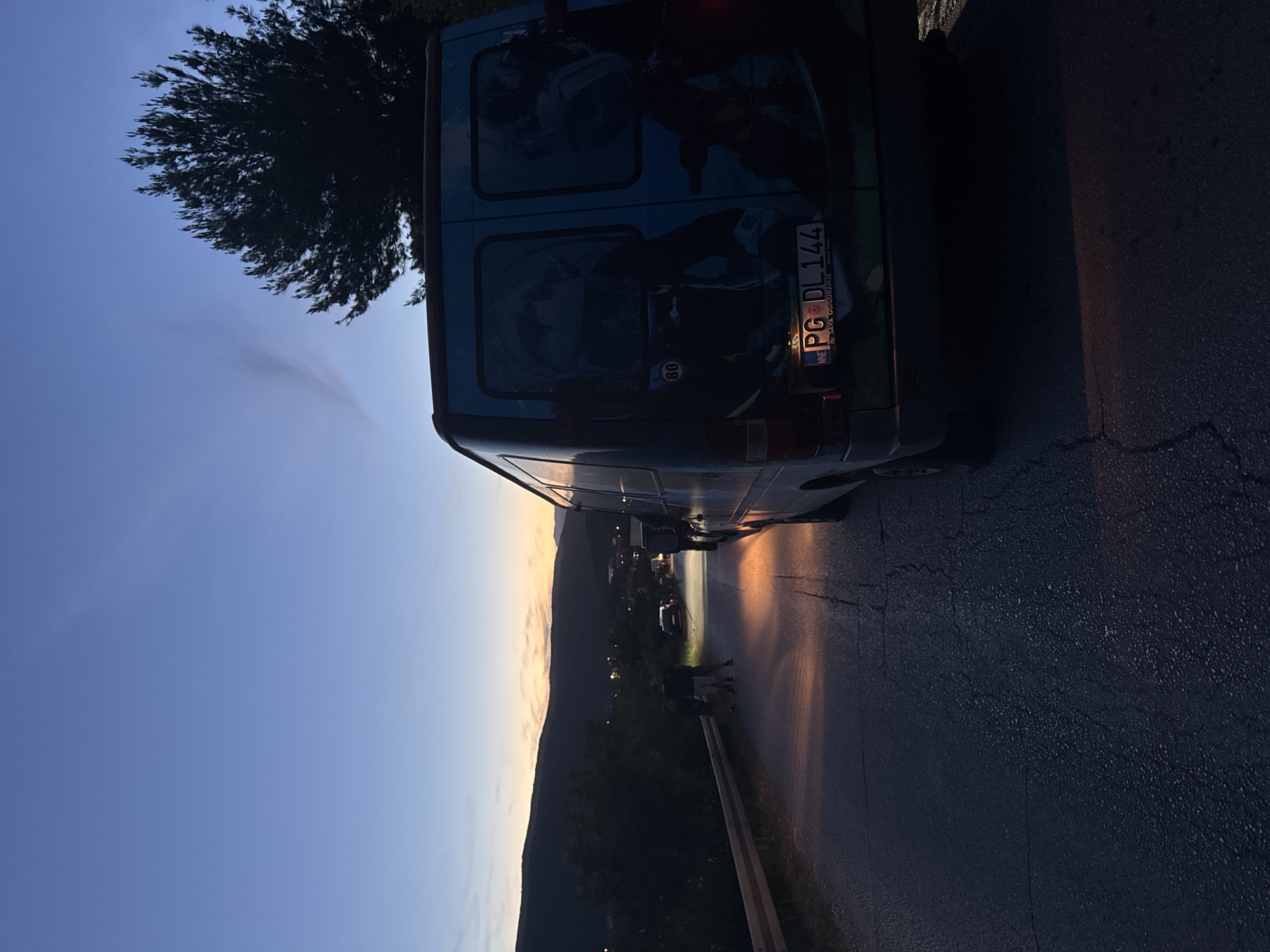 The image depicts a sunset or dusk scene featuring a light blue van parked on the side of an asphalt road, adjacent to a guardrail. The van is custom-painted with graphics of a person on a motorcycle and has "80" in a circle, possibly a speed limit sticker. Its Montenegrin license plate, "PG DL 144", is clearly visible, along with "MNE SAVA OSIGURANJE" below it, indicating its origin from Podgorica.
In the foreground, a dark, dense tree is silhouetted against the sky. The sky displays a gradient from deep blue on the left to warm orange and yellow hues on the right horizon, typical of sunset. Below the horizon, a mountain range extends across the background, with distant lights suggesting a valley or settlement.
Two or three silhouetted figures are standing near the guardrail on the roadside, appearing to be observing the view or waiting. The low sun casts a warm, soft light on portions of the road and the side of the van, creating long shadows and highlighting surface cracks. The overall scene conveys a tranquil moment on a road overlooking a landscape, possibly at the end of a day.