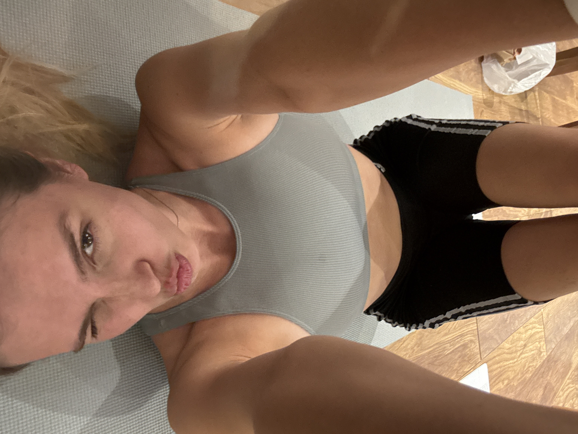 A selfie of a person lying on a grey exercise mat is seen from a first-person perspective. The person is wearing a grey sports bra and black shorts with white stripes. The person's head is tilted slightly towards the camera, and they are making a puckered-lips expression. The mat is situated on a wooden floor. There is a white plastic bag visible in the upper right corner of the image. The location is Unknown, Unknown.