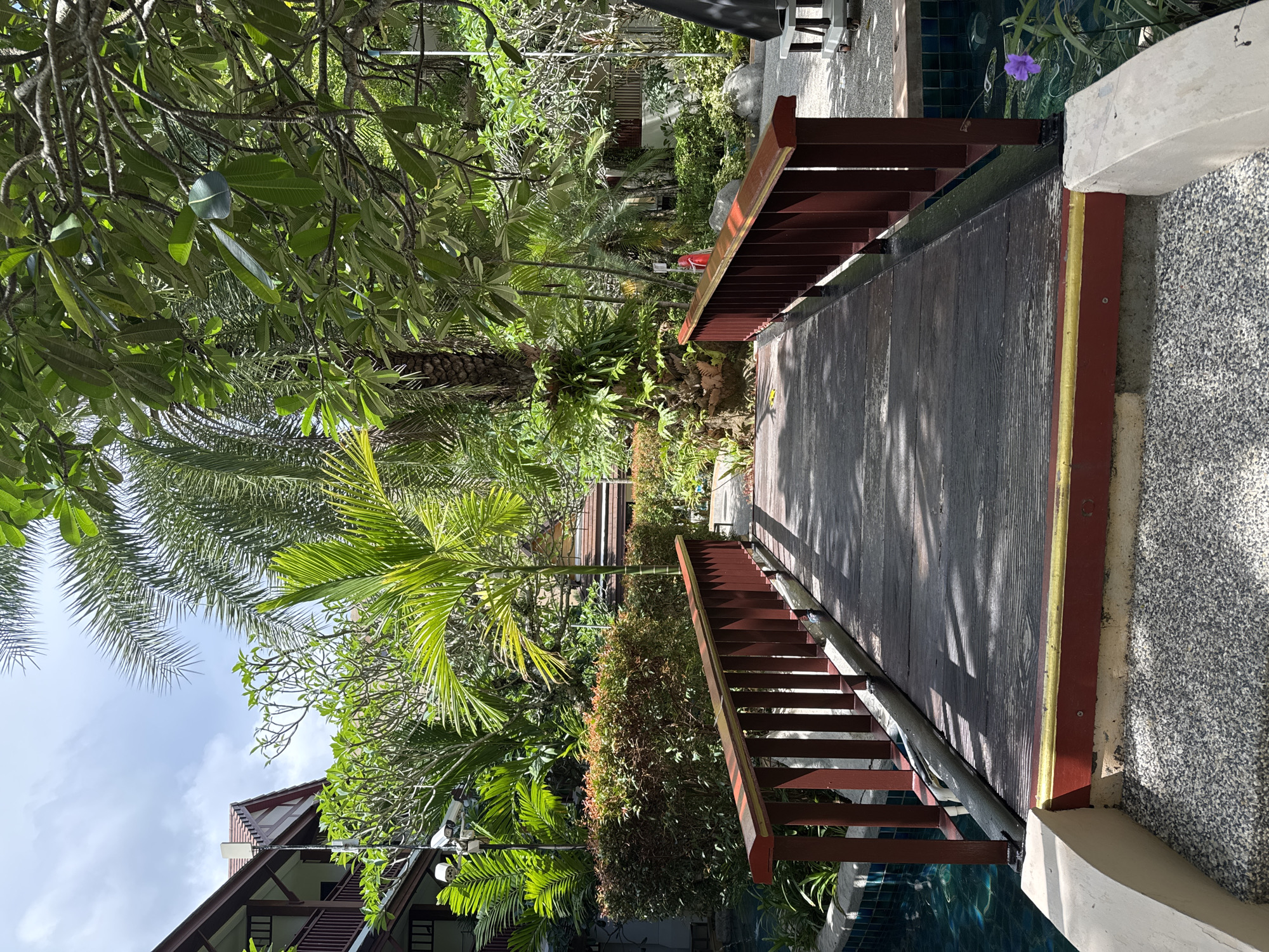 A wooden footbridge with red railings crosses a body of water, likely a decorative pond or shallow canal, in a tropical setting. The bridge has a weathered, dark wood surface with visible grain. Lush green vegetation, including palm trees and other broad-leafed plants, surrounds the bridge and a building with a red-tiled roof and wooden balconies is visible in the background. The area appears to be a resort or a well-maintained garden space. The sky is partly cloudy. A single purple flower is visible in the water to the right of the bridge. The image captures a tranquil outdoor scene in Karon, Thailand.