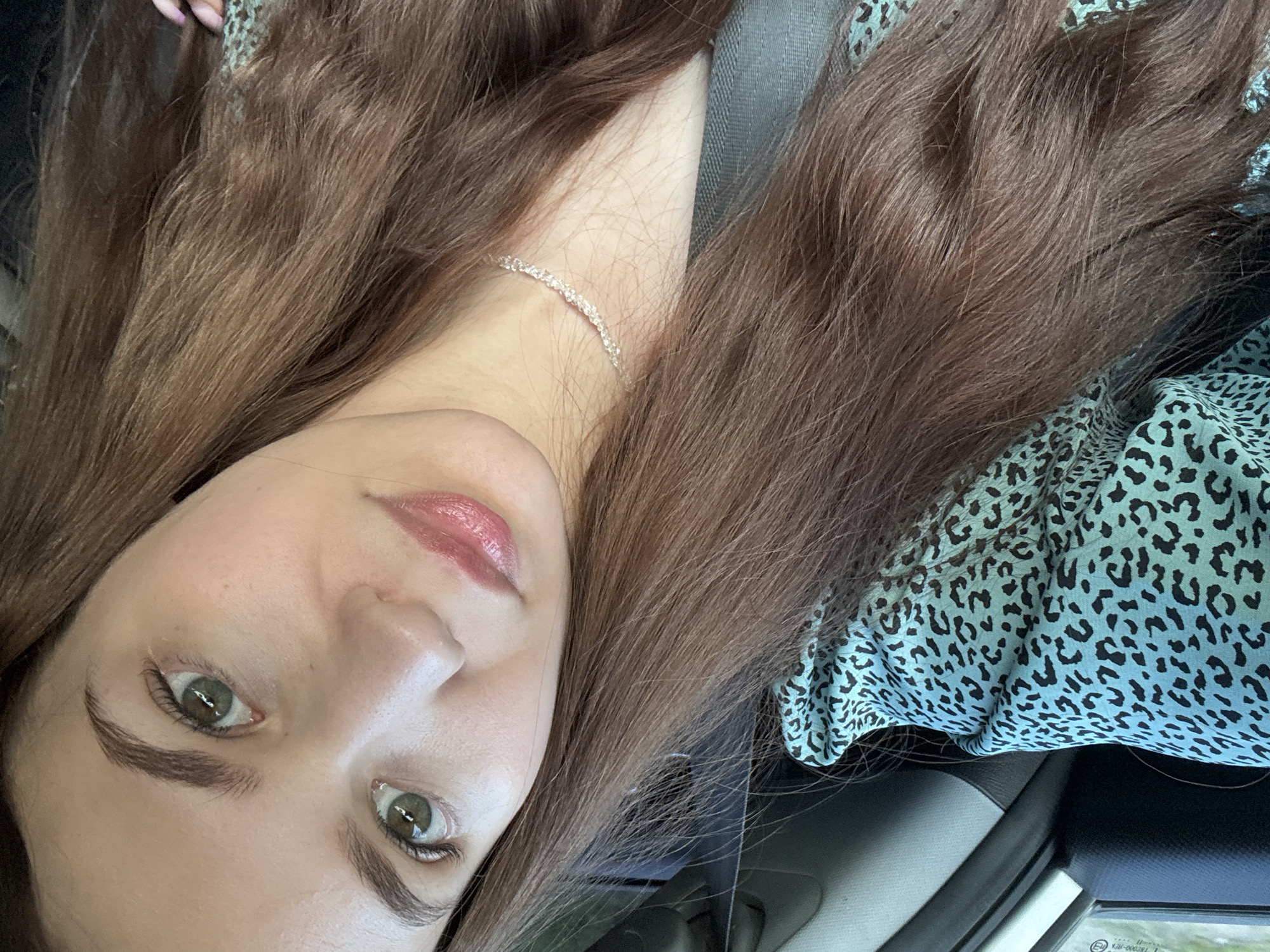 A close-up, angled selfie shows a young woman with long, brown hair, wearing a turquoise and black leopard-print top. She has light skin, green eyes, and is wearing a delicate, sparkly necklace. The woman is inside a vehicle, as indicated by a seatbelt visible across her shoulder and the interior of the car in the background. The setting appears to be daytime, with natural light filtering in from outside the car window. The specific location within the city of Chaikovsky, Russia, cannot be determined from the visual information provided in the image.
