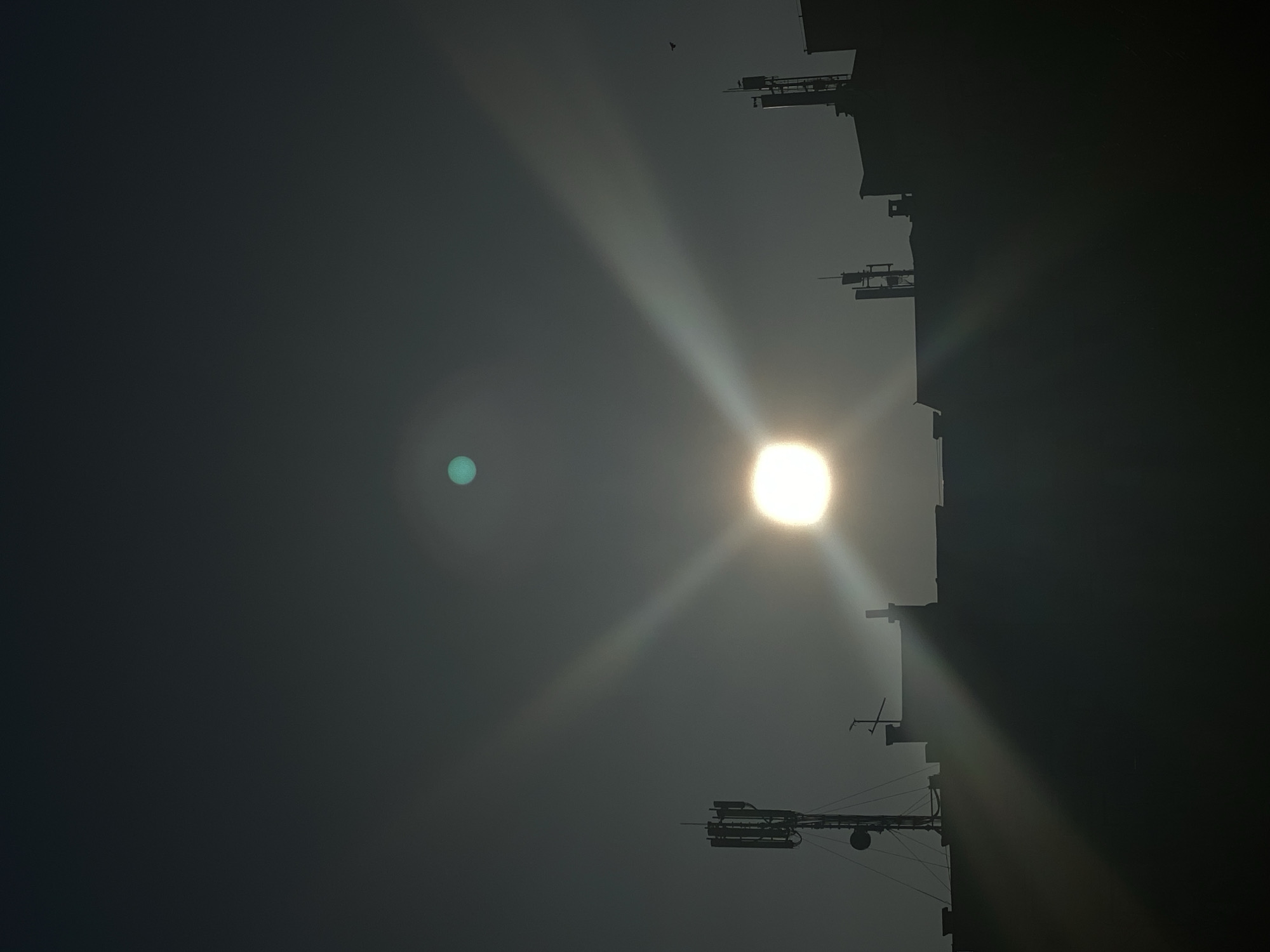 The image captures a stark, upward view, likely taken during the daytime, given the presence of a bright light source (likely the sun) positioned on the right side of the frame. The strong light creates pronounced lens flare and radiant beams that dominate the sky. Adjacent to the light source is the dark silhouette of what appears to be the side of a building, featuring multiple protruding structures resembling antennas or satellite dishes. Higher up in the image, a faint, dark speck, possibly a bird, is visible. A bluish-greenish anomaly exists in the upper-left quadrant of the image.
Due to limited information and lack of landmarks, the precise location within Pleven, Bulgaria cannot be determined solely from this image.