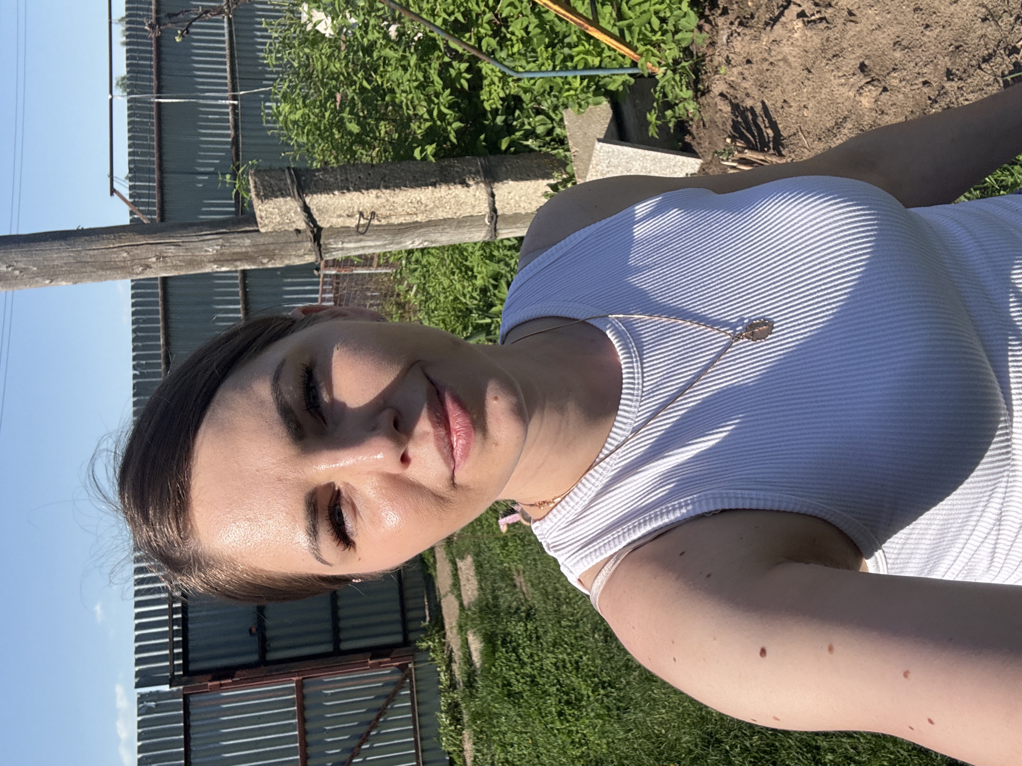 A daytime, medium-angle shot in Razdol'ye, Russia, features a woman in a white, ribbed tank top with a gold necklace. She is positioned in front of a corrugated metal fence, with a wooden post and concrete blocks. Green vegetation and a small patch of dirt are visible in the background. The sky is blue. The woman's right arm is in the foreground, showing moles. The lighting is sunny, creating shadows on her clothing.