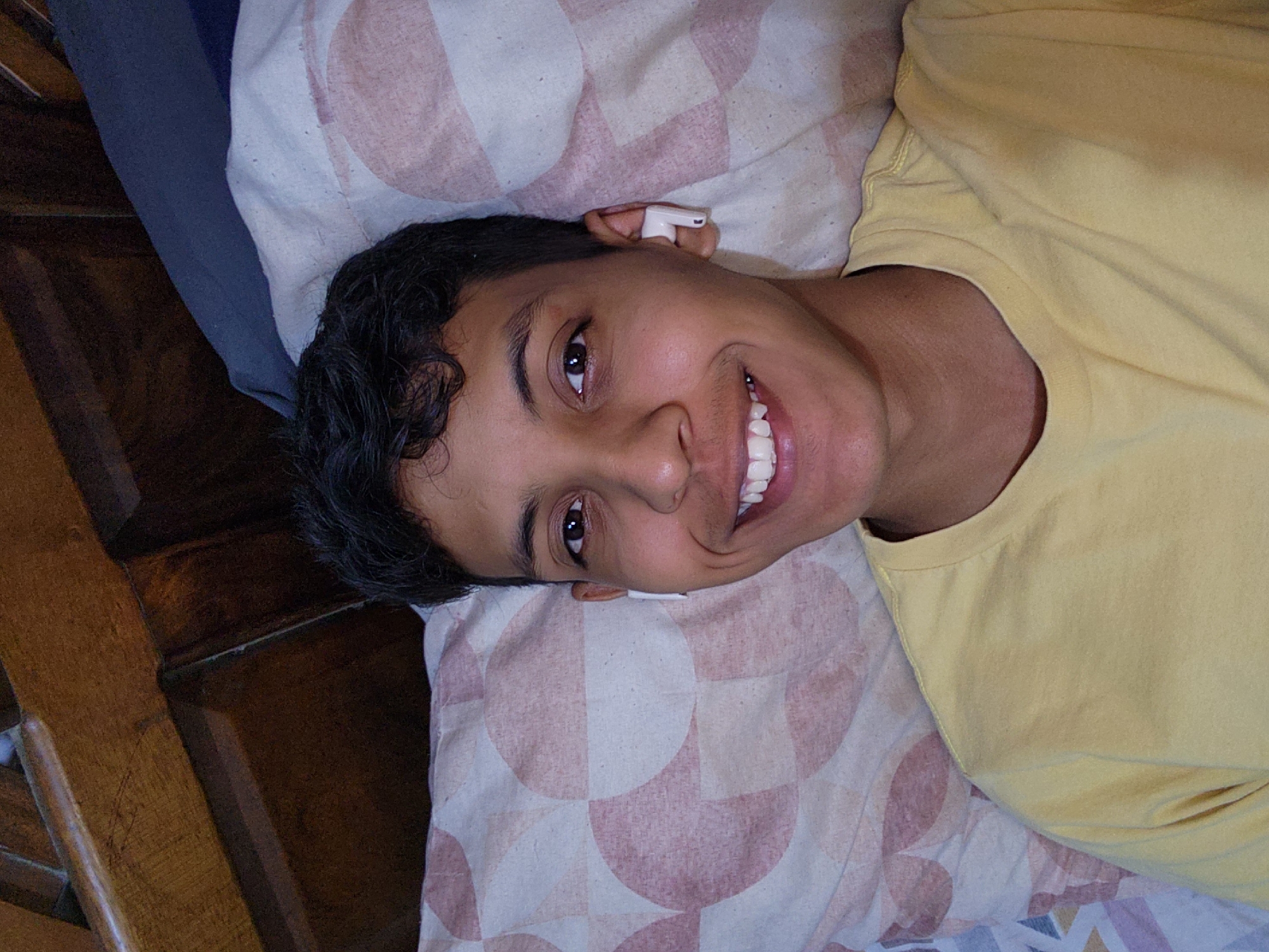 A smiling young man is lying on a patterned pillow in what appears to be an indoor setting. He is wearing a yellow shirt and has an earbud in his left ear. A portion of what may be wooden furniture is visible on the left side of the image. The location in Tinaquillo, Venezuela is not discernible from the image.
