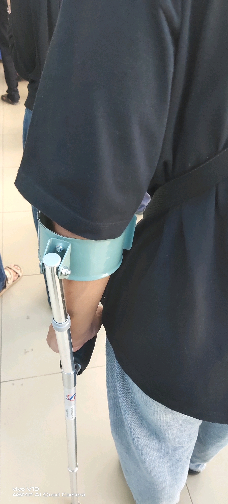 This image shows a close-up of a person's arm and lower body, supporting themselves with a forearm crutch. The crutch has a teal-colored cuff that encircles the forearm, secured by metal brackets and bolts. The shaft of the crutch is made of silver-colored metal. The person is wearing a black t-shirt and light blue jeans. In the background, other individuals are partially visible, and the floor appears to be tiled. The overall setting suggests an indoor public space. The image also contains a watermark in the lower left corner: "vivo V19 48MP AI Quad Camera". The location of Pekanbaru, Indonesia is not discernible from the image itself.