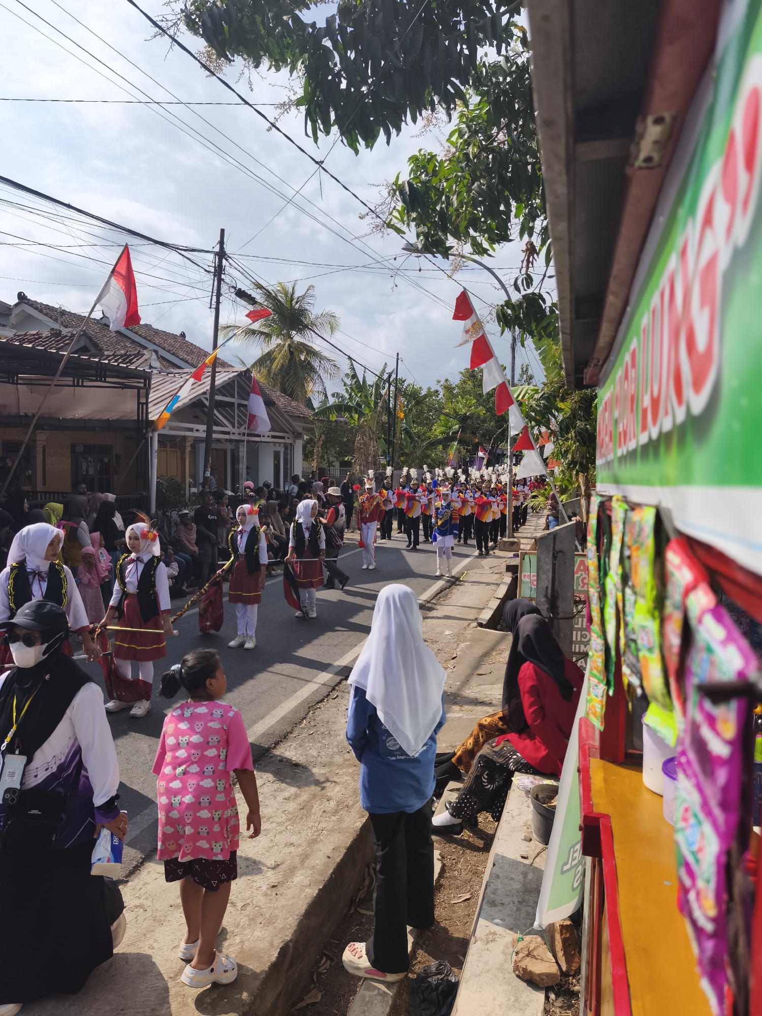In Kutoarjo, Indonesia, a street parade is in progress under a partly cloudy sky. A marching band, dressed in colorful uniforms with gold accents and hats, proceeds down the street. Several participants, some carrying batons, are visible in formation. The street is lined with spectators, including women wearing hijabs and a young girl in a pink dress with owl prints. Houses with red and white flags, symbolizing Indonesian independence, are visible along the sides of the street. A stall with green and red signage is set up on the right side of the road, displaying various goods. Power lines and trees with green foliage also feature in the scene.