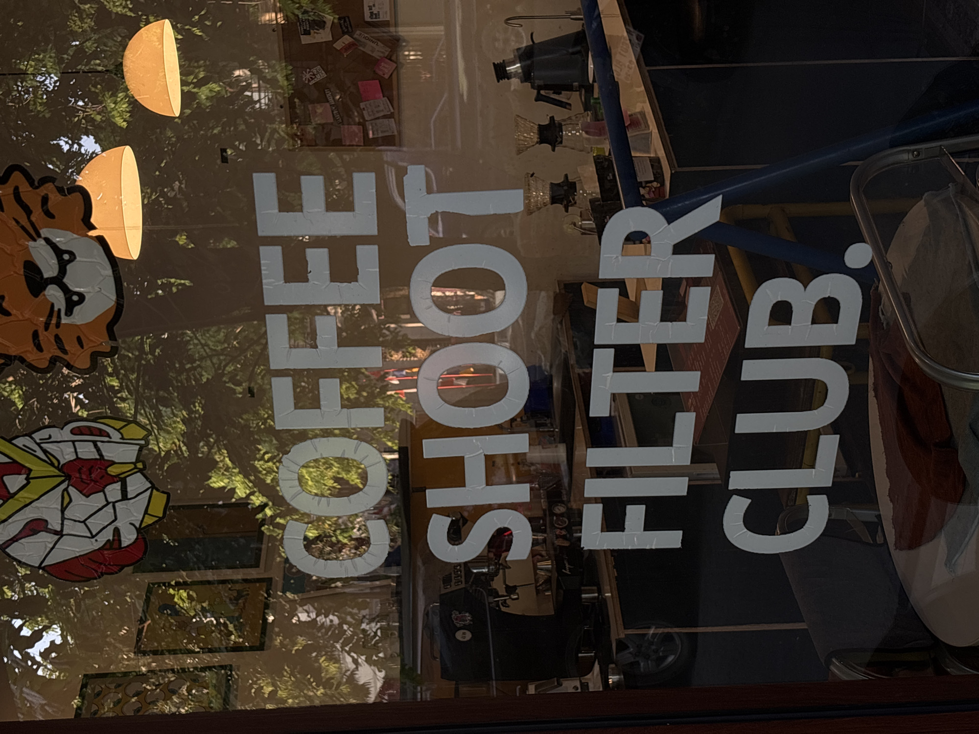 The image shows a window with white lettering that reads "COFFEE SHOOT FILTER CLUB." The text is applied to the exterior of the glass, with some wear and tear visible on the letters. Through the window, an interior of a coffee shop is visible.
On the left side of the window, a black espresso machine is prominent. Behind it, there are shelves with various items, including what appears to be posters or framed artwork.
The upper part of the window features cartoonish decorations: on the left is a stylized robot head, and on the right is the face of a tiger with a grumpy expression. Two orange dome-shaped lamps hang from the ceiling in the background.
In the middle and lower sections of the window, more of the coffee shop interior is visible. There are coffee brewing equipment, including pour-over coffee makers and a kettle, on a counter.
The reflection on the window shows trees and foliage, suggesting an outdoor setting with greenery. There are also reflections of street lights or lamps.
The text on the window and the overall aesthetic suggest this is a coffee shop. Based on external knowledge, this establishment, "Coffee Shoot Filter Club," is located on Jalan MT Haryono in Semarang, Indonesia. The time of day appears to be late afternoon or early evening, given the lighting and shadows. No people are clearly visible in the image, only reflections and objects within the shop.