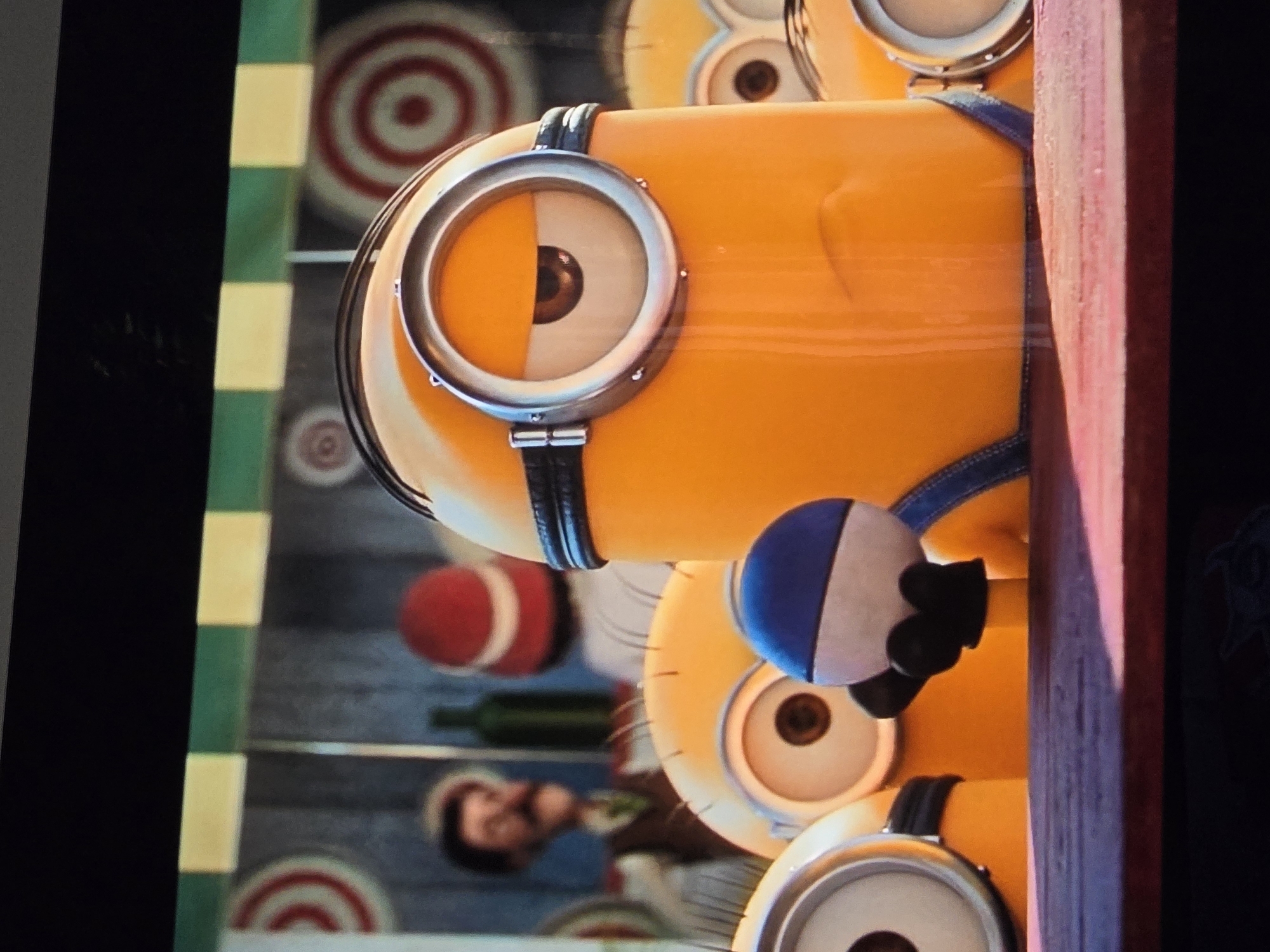 The image shows a close-up of several Minions, popular animated characters, in what appears to be a carnival or fairground setting. In the foreground, a large Minion with one eye is prominently featured, wearing blue overalls. It is holding a blue and white striped ball with its gloved hands. Behind this Minion, other Minions are partially visible, also with their characteristic goggles and yellow bodies. In the background, a man with a mustache wearing a brown vest and a green tie is visible, as well as a red hat with a white stripe, and green bottles. The backdrop appears to be a structure with targets resembling dartboards. The lighting suggests it is daytime, possibly indoors or under a covered area due to the stripes of green and yellow across the top, which could be a tent or awning.