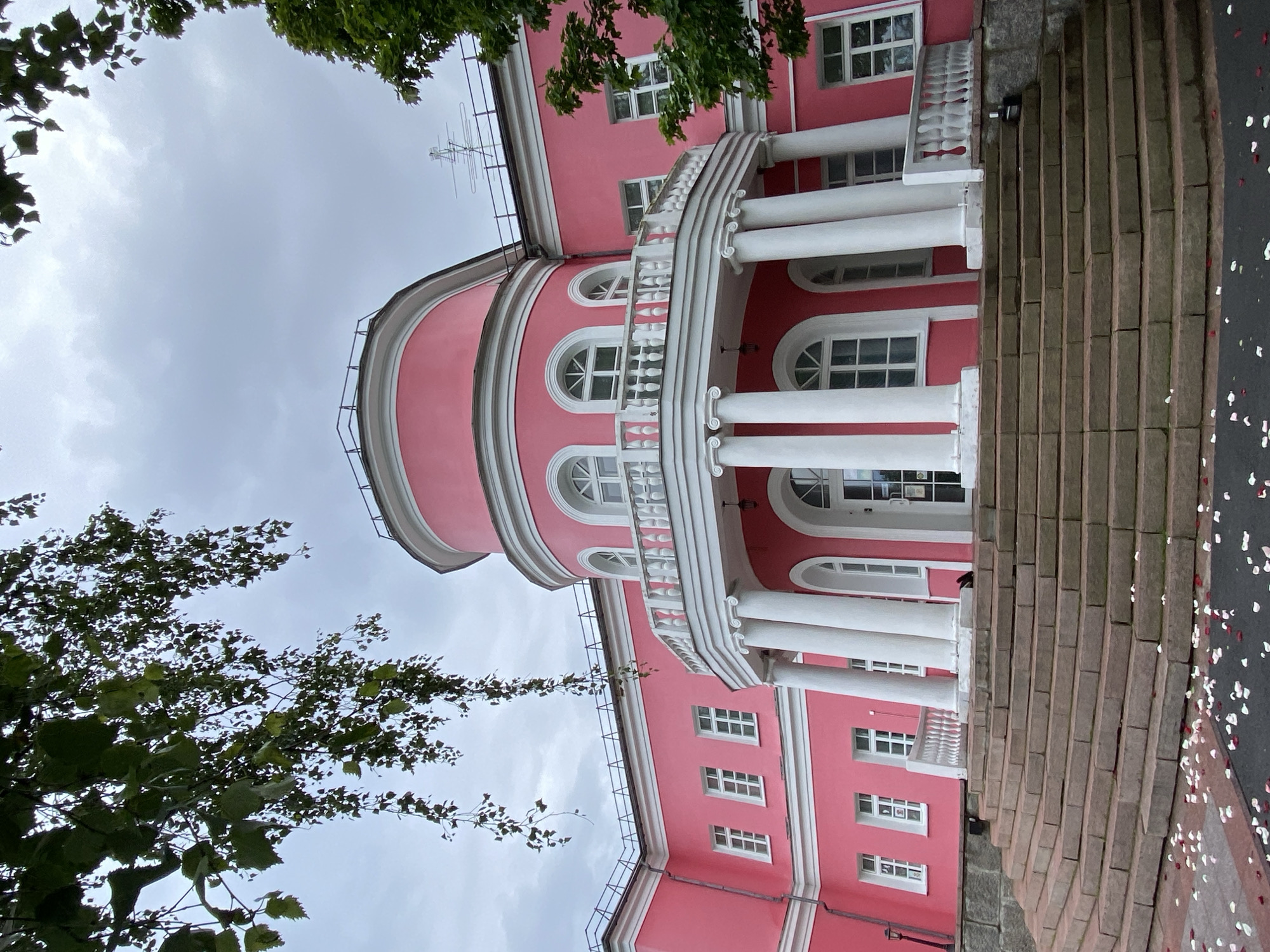 The image shows the North Caucasian Railway Museum in Rostov-on-Don, Russia, specifically its striking pink building. The building features a rounded portico with white columns, a white balustrade, and arched windows. A wide, curved stone staircase leads up to the entrance. The facade is painted a vibrant pink, contrasting with the white architectural details. Lush green trees frame the building, and the sky is overcast with clouds. The foreground of the staircase and the surrounding pavement are scattered with white and red flower petals. There are no people visible in the image.