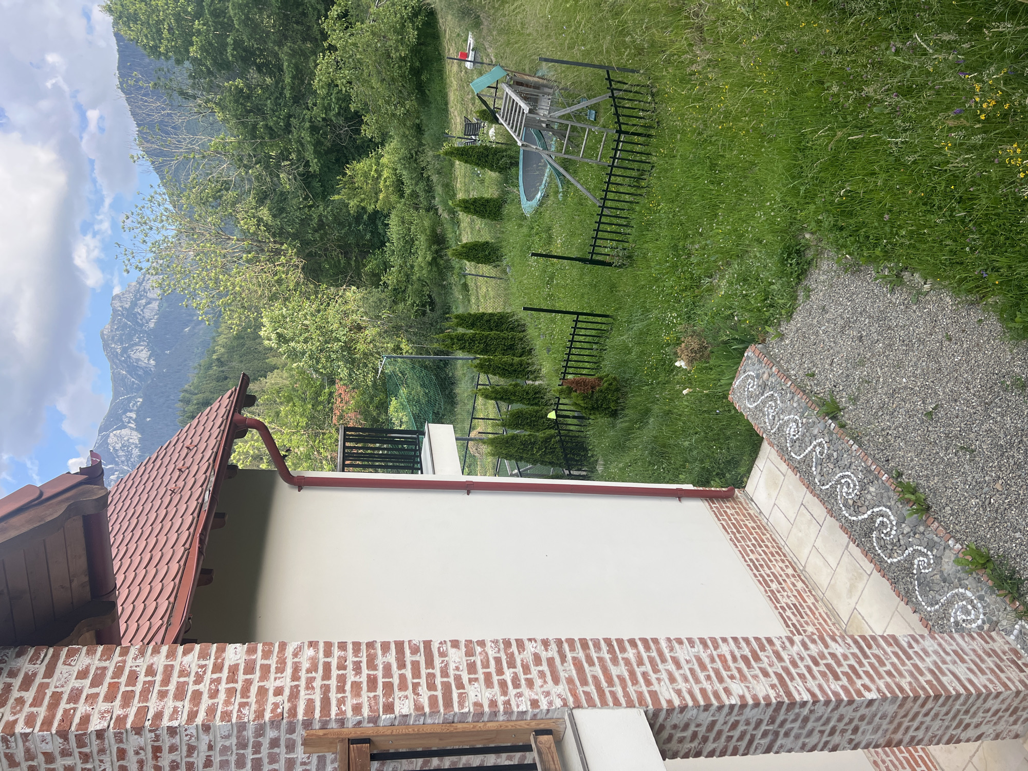 The image shows a side view of a building with a red tiled roof and a brick chimney. The building has a white exterior wall. In front of the building, there is a garden with a trampoline and a wooden play structure. The garden is surrounded by a black metal fence and several coniferous trees. The background features a dense forest with mountains visible in the distance. The sky is partly cloudy. The scene appears to be a residential property in a rural or mountainous area.