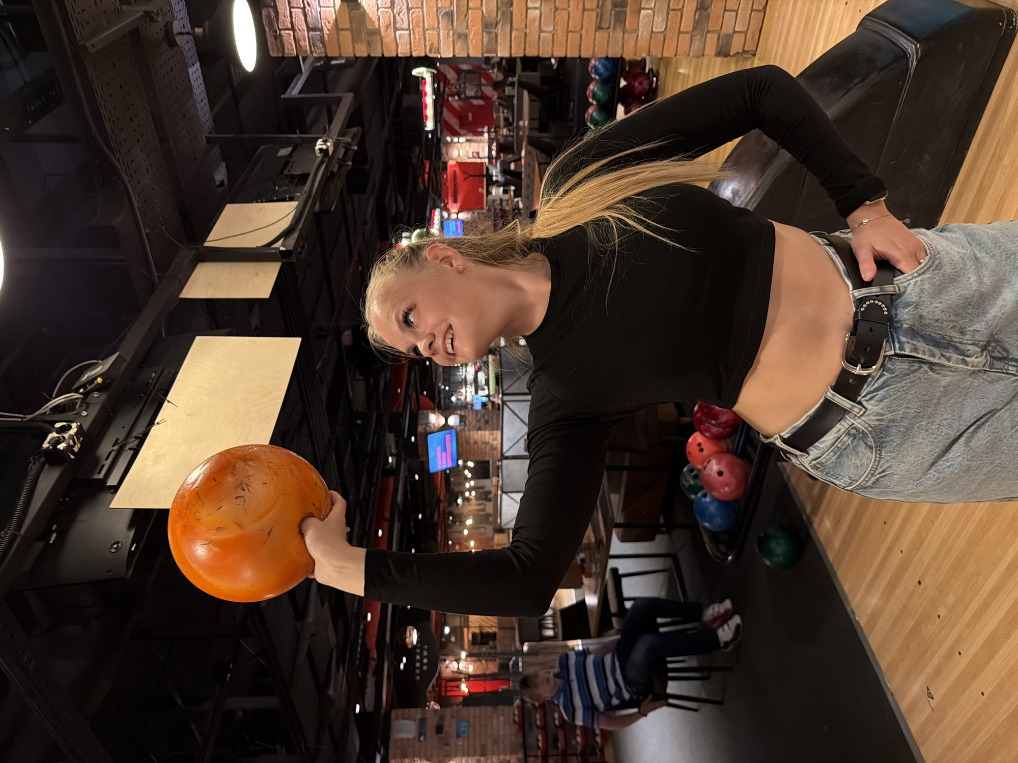 A young woman with blonde hair in a ponytail, wearing a black long-sleeved crop top and light-wash jeans, holds an orange bowling ball above her head with her right hand in a bowling alley. She is looking upwards and to her right with a smile. In the background, a man wearing a blue and white striped shirt and dark pants sits in a chair. Several bowling balls are visible on a rack in the middle ground. The environment includes brick walls, wooden flooring, and overhead lighting. The image was taken in Izhevsk, Russia.