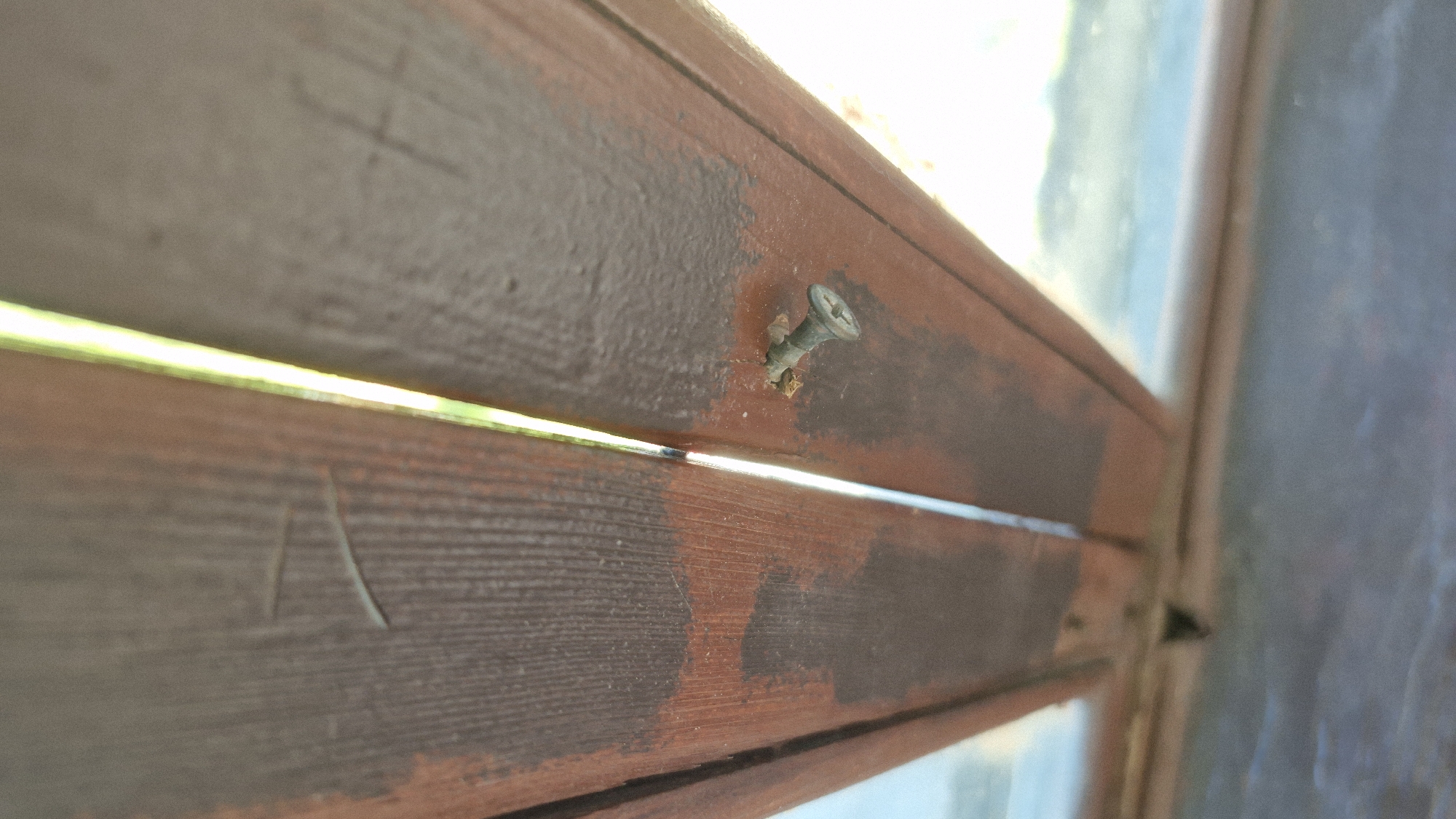 The image shows a close-up of a brown painted wooden frame, possibly a window or part of a structure, with a screw partially embedded in the wood. The wood has a textured surface and is composed of horizontal planks. The screw is slightly angled, and there are wood shavings visible around it. The image is taken at an angle, focusing on the screw and the wood. Beyond the frame, there is a blurred view of what appears to be an exterior, with sky and/or other structures visible.