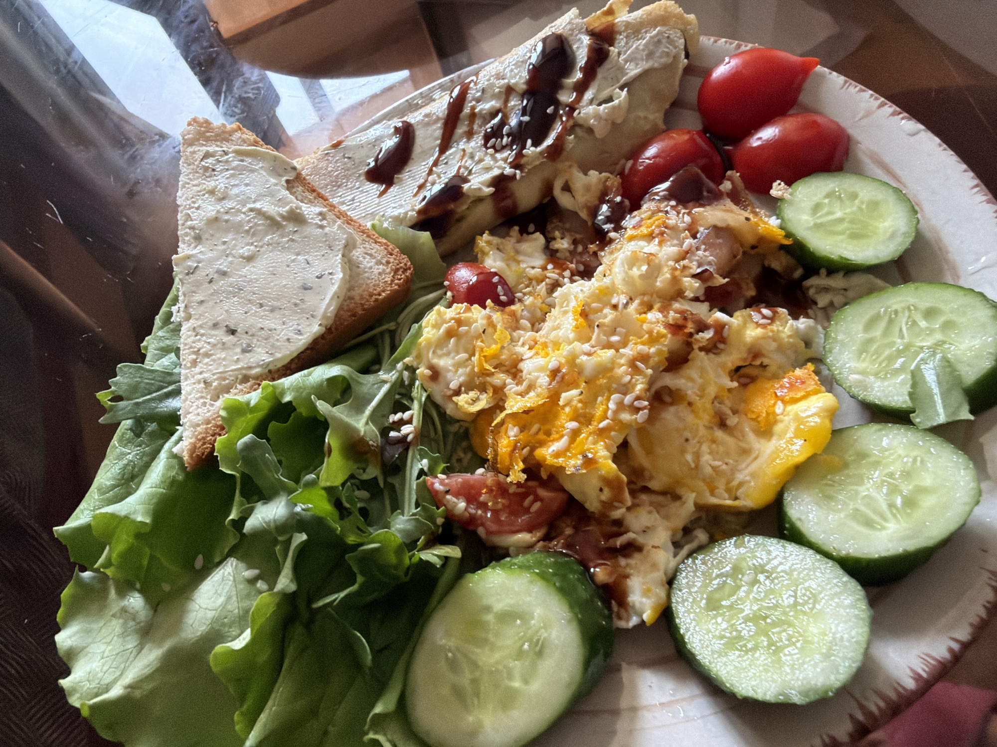 A plate of food is on a table. The plate contains scrambled eggs, a sandwich, lettuce, cherry tomatoes, and cucumber slices. The food appears to be a breakfast or brunch meal. The lighting is natural and comes from an unseen window. The table appears to be made of wood. The scene is indoors, likely in a home. The location is not specified in the image.
