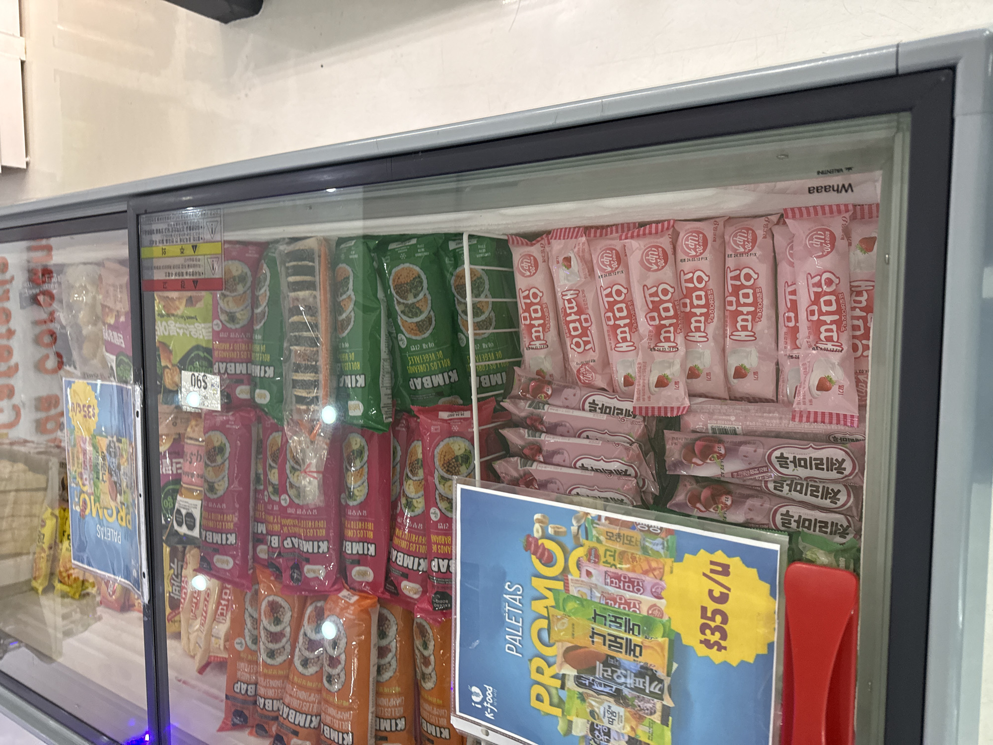 The image shows a refrigerated display case in what appears to be a store in San Andrés Cholula, Mexico. The refrigerator is stocked with various frozen Korean snacks and treats. The front of the display case has several promotional signs, including one that advertises paletas (popsicles) at a price of $35 pesos each. There are various brands and flavors of frozen treats visible, including Korean kimbap, and a number of Korean popsicles. The products are neatly arranged inside the refrigerator. The red handle of the display case is visible on the right side. The general setting indicates a small retail environment where these Korean frozen foods are sold.