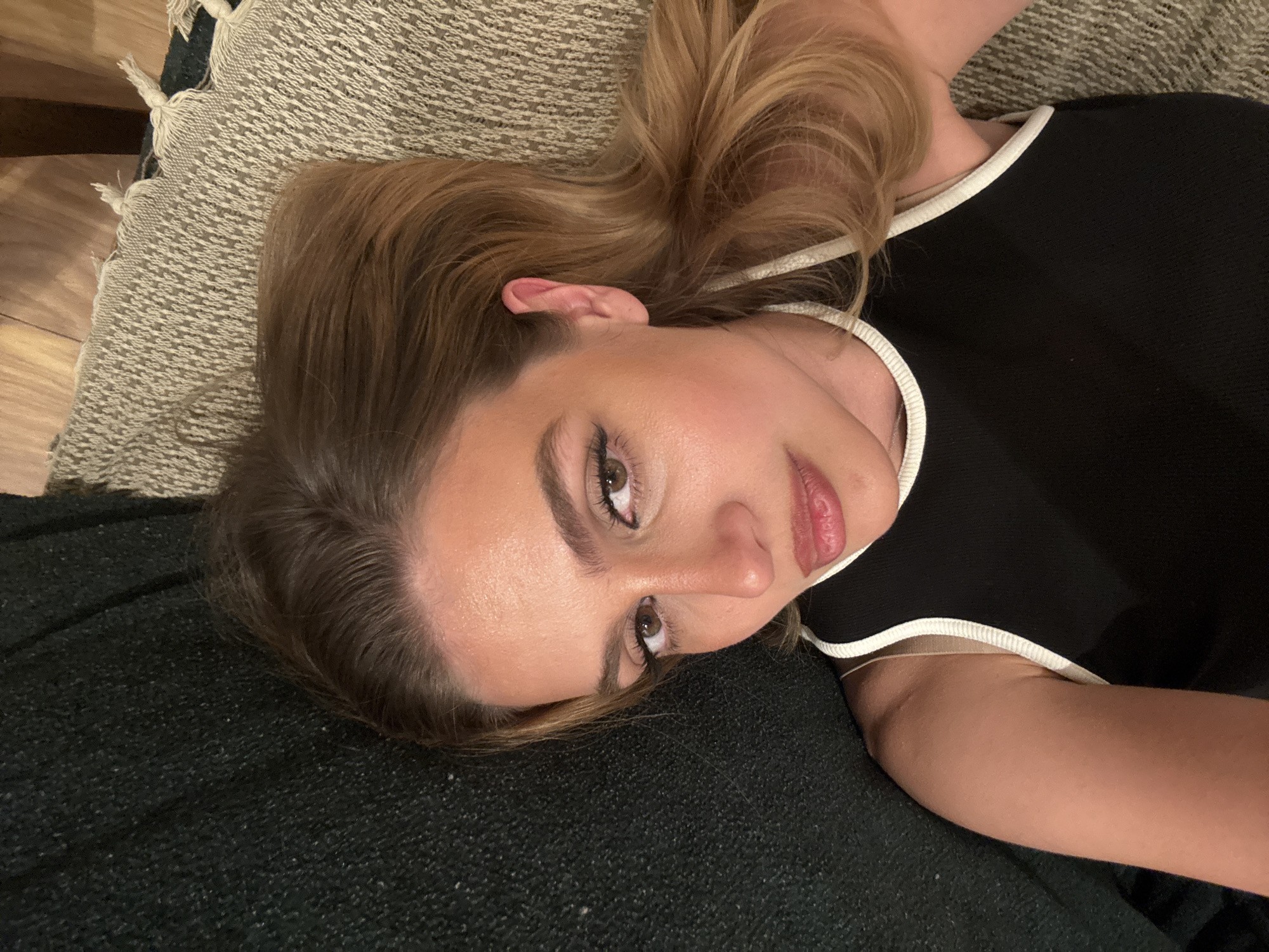 A portrait-style shot shows a young woman lying down indoors, looking towards the camera with a direct, relaxed gaze. Her head is tilted to her right. She has light brown hair with blonde highlights, which is spread out on a textured, dark gray or black fabric, likely a blanket or throw. Her skin is fair, and she is wearing makeup, including visible eyeliner, defined eyebrows, and light pink lipstick. She is dressed in a black top with white trim along the neckline and sleeves.
Next to her head, a lighter, patterned woven fabric, possibly a rug or another throw, is visible, showing a distinct geometric pattern. A small portion of a wooden surface, likely a floor or piece of furniture, can be seen in the upper left background. The overall lighting suggests an indoor setting, possibly under ambient or artificial light. No text is visible in the image.