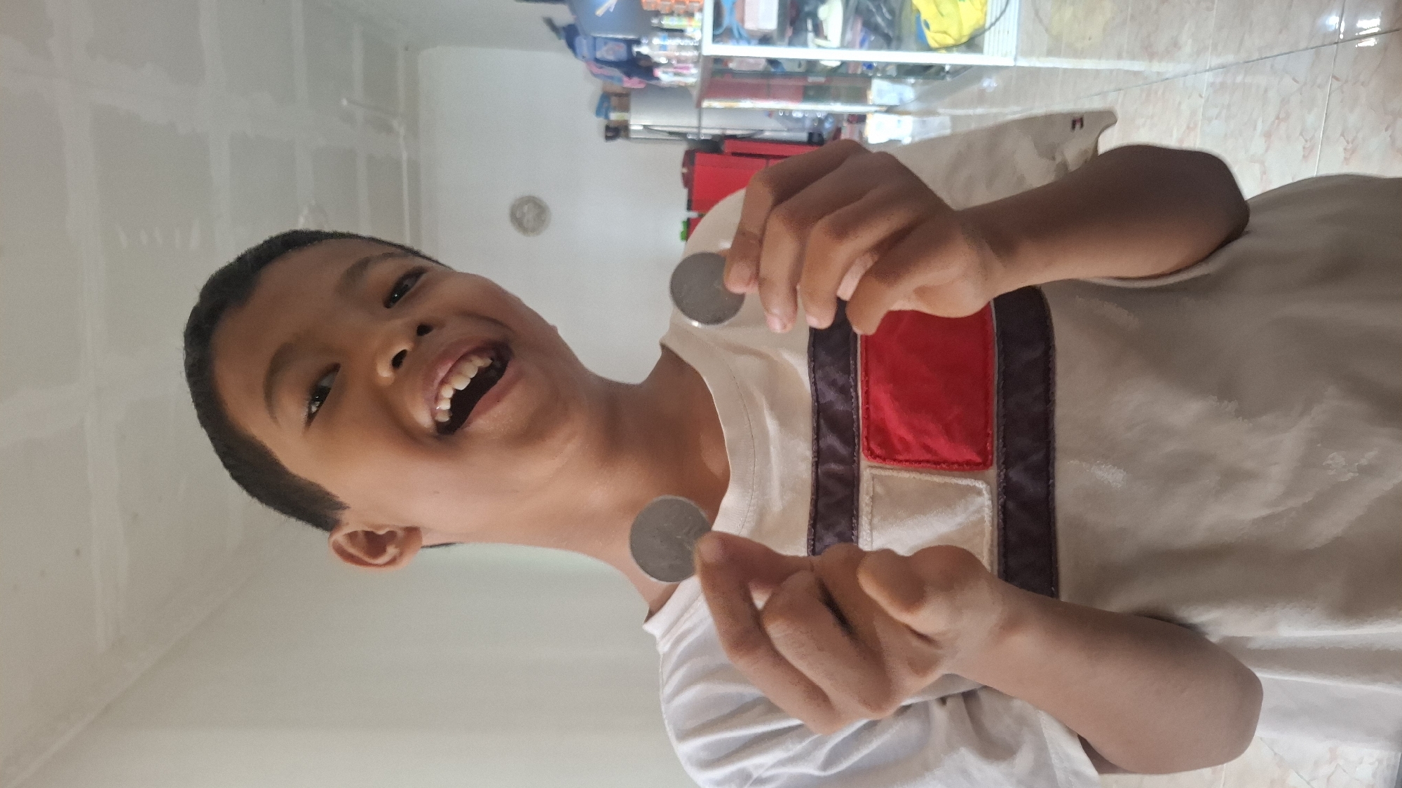 A boy is holding two coins, one in each hand, up to the camera. He is smiling widely with his mouth open. He is wearing a white t-shirt with red and dark blue accents on the chest. The background appears to be an indoor setting, possibly a living room or hallway, with tiled flooring and a shelf unit in the distance. The lighting suggests it is daytime.