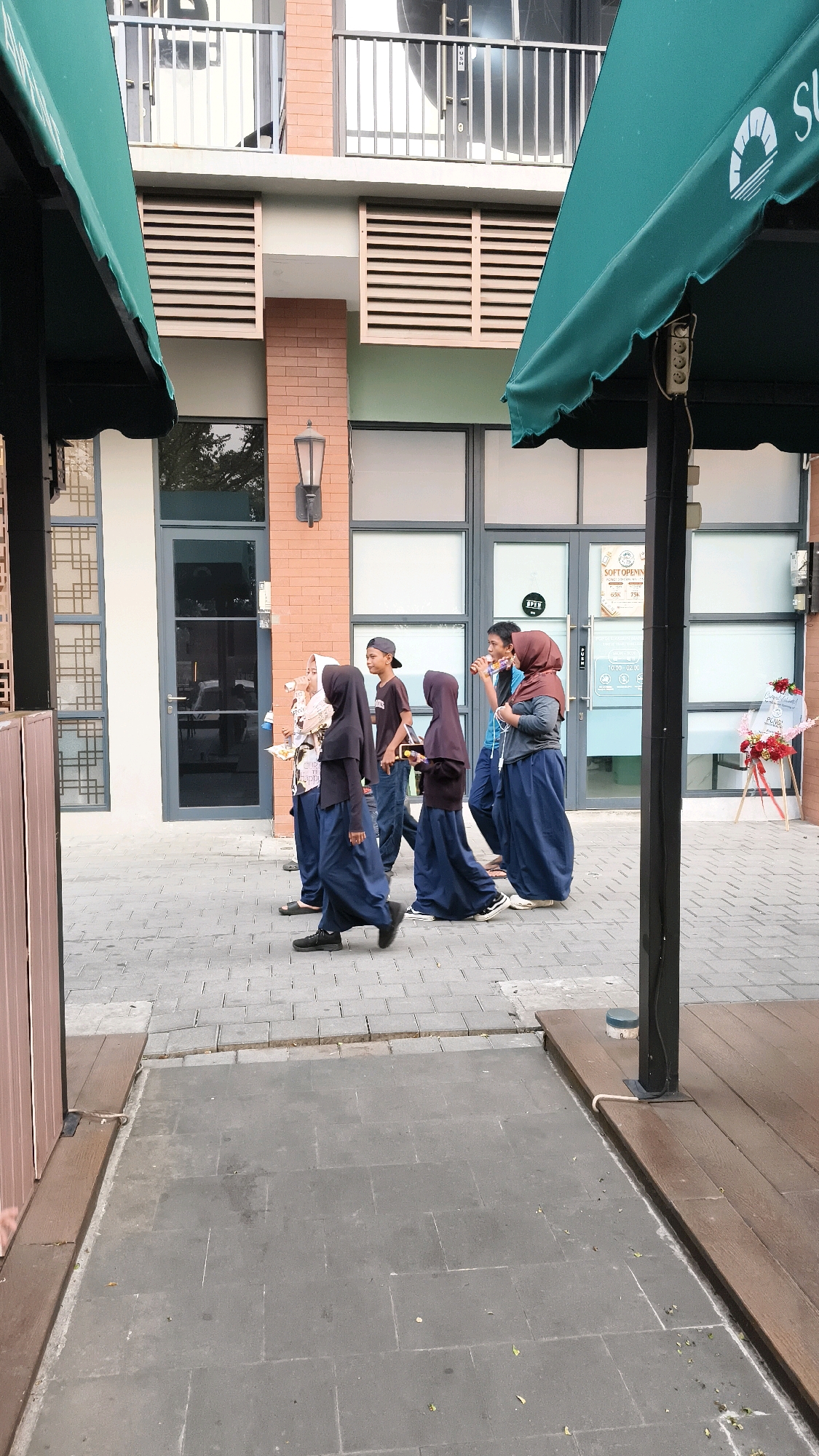 In West Jakarta, Indonesia, a group of five individuals, appearing to be students, walks along a paved walkway. The group consists of three females wearing dark hijabs and long dark skirts, and two males. One male wears a dark t-shirt and a baseball cap, while the other wears a light blue t-shirt. The individuals are walking from left to right in the frame. The background features a modern building with brick accents, large glass windows, and outdoor awnings. The walkway is paved with interlocking grey bricks. To the right of the group, a wooden deck area is visible with a dark vertical pole. In the far right of the image, a red and pink floral arrangement is displayed on an easel. The scene appears to be an outdoor area in front of a commercial establishment.