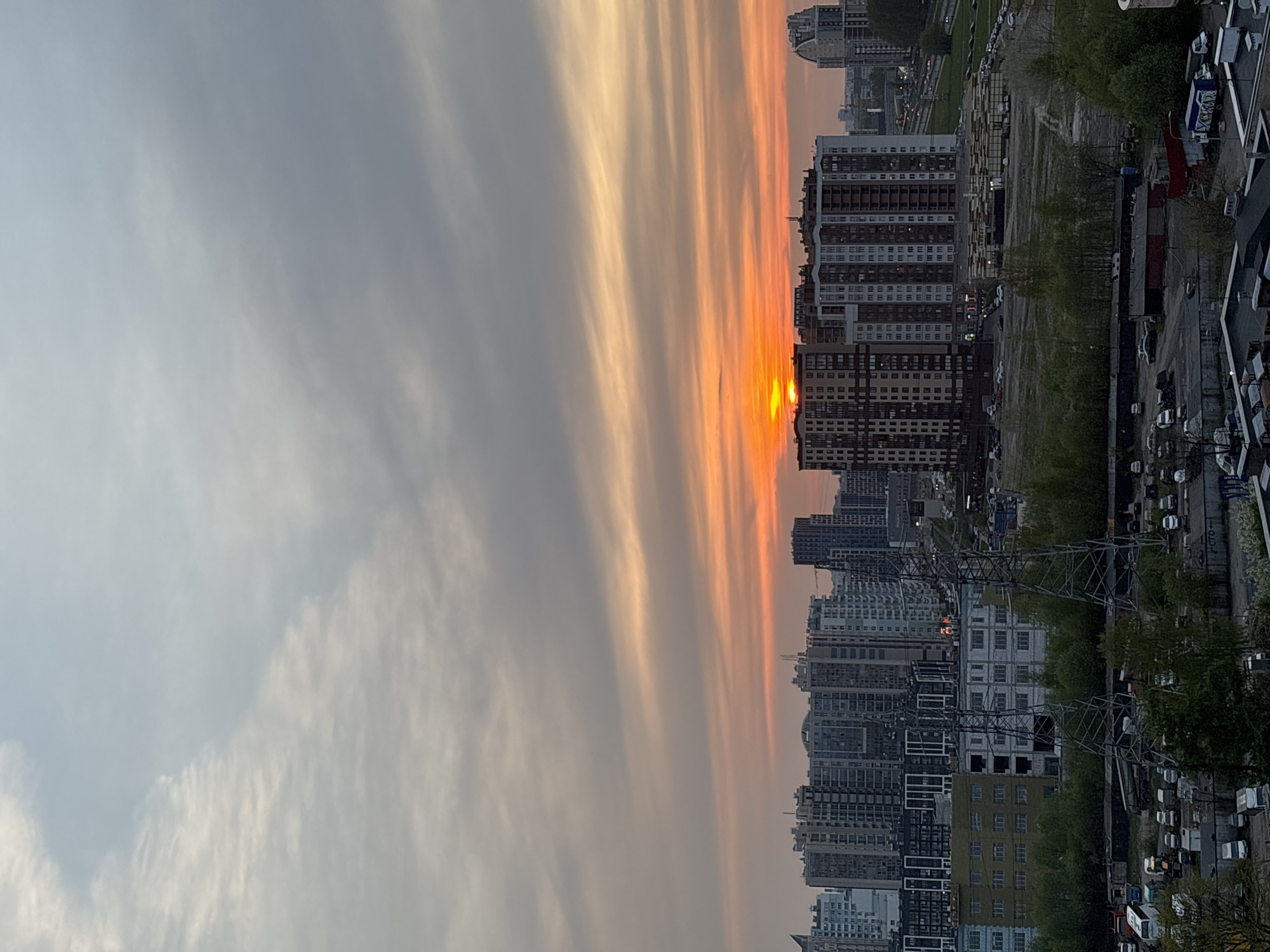 The image depicts a cityscape at sunrise or sunset. Tall buildings, mostly of a modern architectural style, dominate the view, with some appearing to obscure the sun on the horizon. The sky is filled with elongated cloud formations colored in orange and gray tones. In the foreground, there is evidence of urban infrastructure, including power lines, roads, and parked cars. The location is an unidentified city.