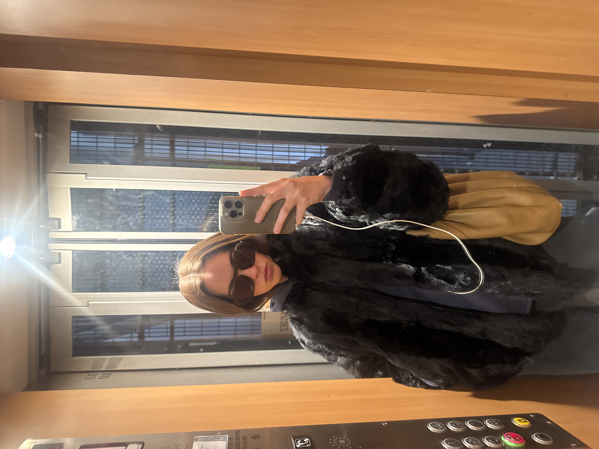 A woman is taking a selfie in an elevator. She is wearing a black fur coat, sunglasses, and holding a phone. The elevator has wood paneling, a window with blinds, and a control panel with buttons. Outside the window, a cityscape with buildings and the sky is visible. The elevator appears to be moving upward. There is no other information in the image to confirm this is the city of Paris, France.