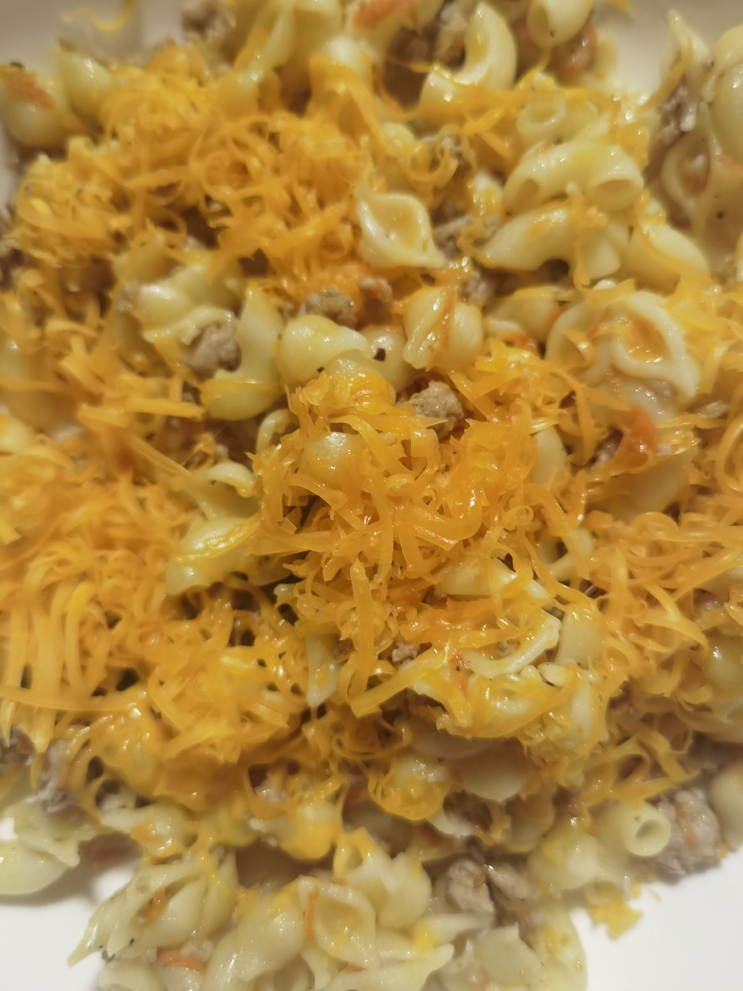 This image depicts a close-up view of a prepared food dish, located in the city of Chaikovsky, Russia. The primary subjects are elbow macaroni pasta, light brown ground meat, and shredded orange cheese. The pasta, which is light cream to yellow in color, forms the base of the dish, appearing to be coated in a light, possibly creamy, sauce. Interspersed among the pasta are numerous small, irregular fragments of cooked ground meat. A substantial quantity of thinly shredded, bright orange-yellow cheese is generously layered over the entire surface of the dish, with some portions exhibiting a melted texture. Small, indistinct orange specks are visible amongst the pasta and meat. The food is contained within a white or light-colored dish, with portions of its rim visible along the edges of the frame. The scene is brightly illuminated, highlighting the textures and colors of the food components.