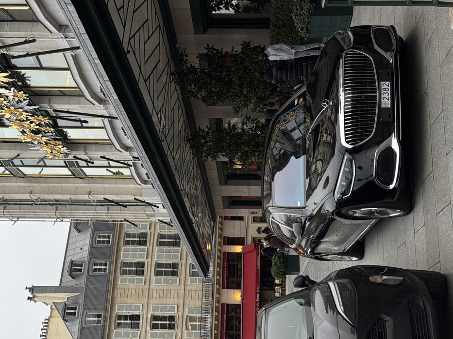 Two black cars are parked on a city street next to a building with ornate architectural details and gold embellishments. The building's windows are visible, as are the rooftops of other buildings across the street. The cars are parked in front of a storefront with a red awning. People are milling around in the background. The angle suggests the photo was taken from a higher vantage point, rotated, showing the scene from an unusual perspective.