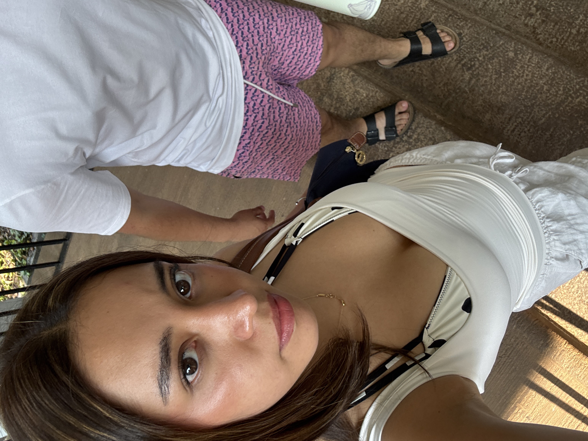 A woman is taking a selfie with another person standing behind her. The woman has brown hair, brown eyes, and is wearing a white dress over a black and white top. The person behind her is wearing a white t-shirt and pink shorts with black sandals. They are standing on a set of stairs. Based on the presence of what appears to be bougainvillea, the image may have been taken outdoors in Cocoyoc, Mexico.