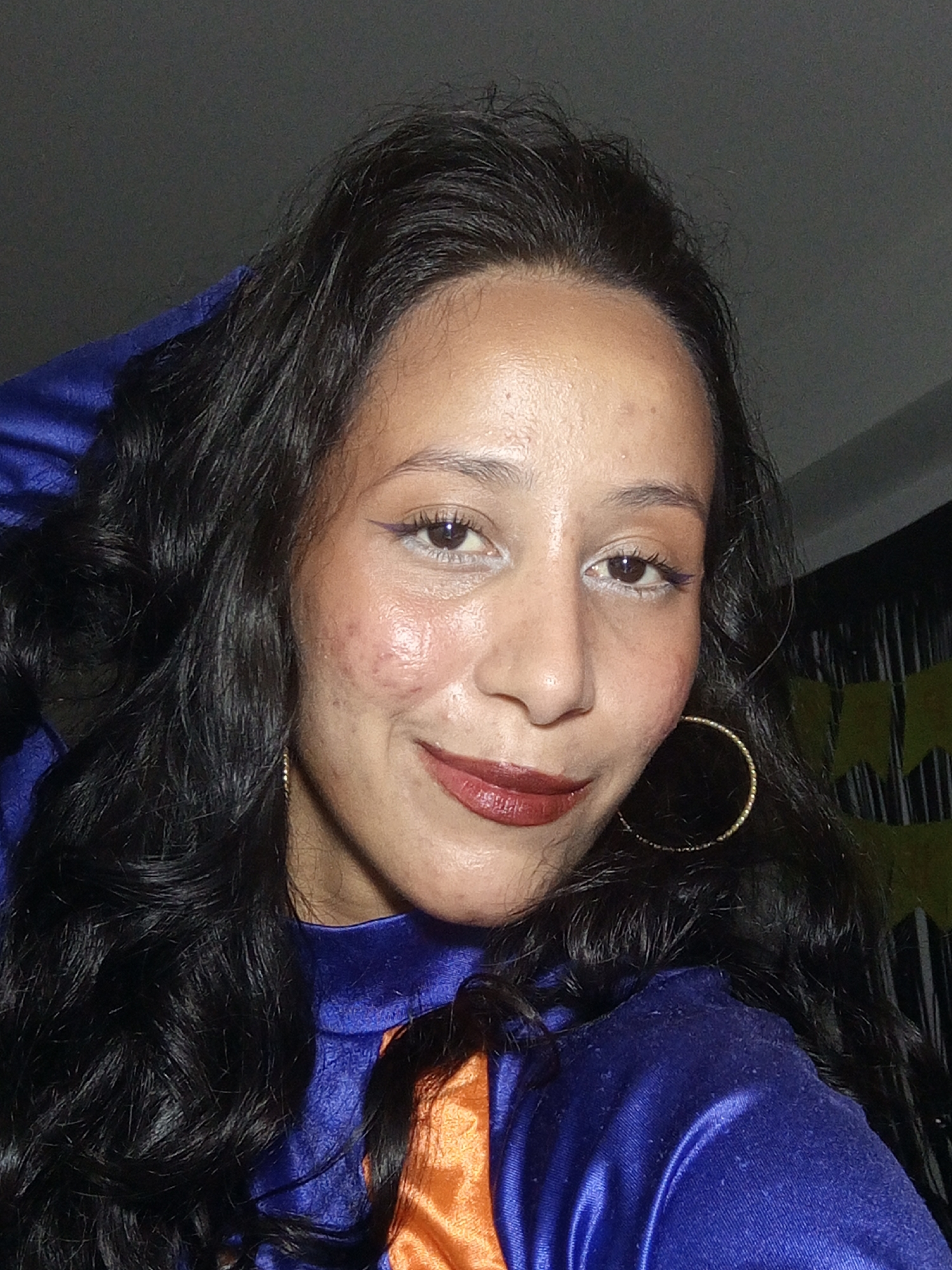 A medium-closeup shot of a woman with dark, wavy hair posing for a picture. She wears a blue and orange outfit with large gold hoop earrings. She has fair skin, dark eyes with light eyeshadow and eyeliner, and wears dark lipstick. Her hand is behind her head, slightly lifting her hair. The background is dark with what appears to be a striped curtain and some yellow decorations. The location is not discernible from the image.