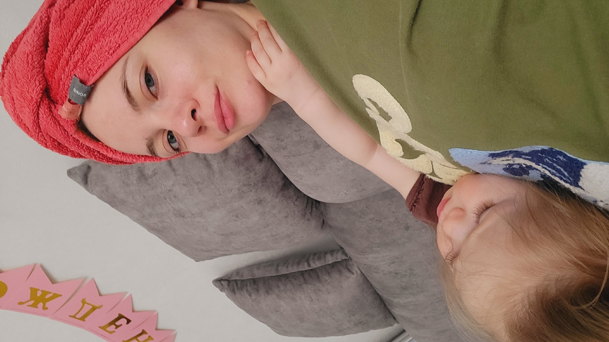 The scene depicts a woman and a child lying on a grey sofa, seemingly indoors. The woman, wearing a red towel wrapped around her head, has light skin and blue eyes. The child, with blonde hair, is lying close to the woman, their arm slightly raised. Both individuals appear relaxed. The woman is lying on several grey pillows.
The woman is wearing an olive green shirt or dress with a pale design on the front. A pink and yellow banner or garland is visible in the lower left corner of the image.
There is no contextual data in the image suggesting the location is Chaikovsky, Russia.