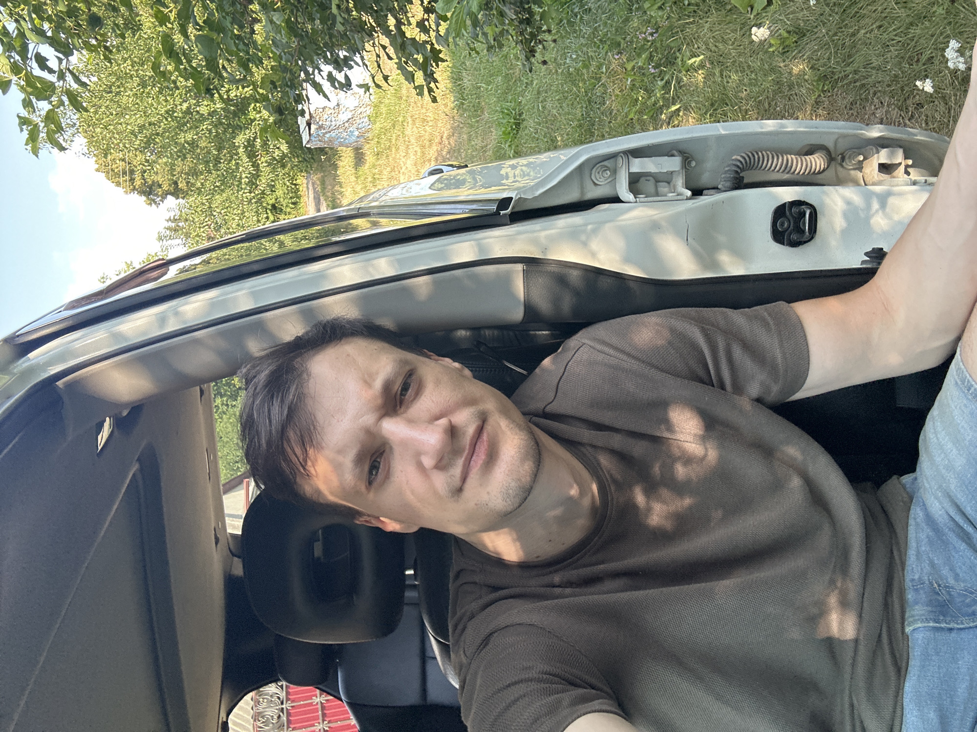 A man is sitting in the driver's seat of a light-colored car. The car door is open. The man is wearing a brown t-shirt and blue jeans. He is looking at the camera and has a neutral expression. Sunlight is casting shadows on his face and shirt. The background is blurred, showing green trees and grass, suggesting an outdoor setting. A small, brightly colored structure is visible in the distance. The scene appears to be in the vicinity of Shatava, Ukraine.