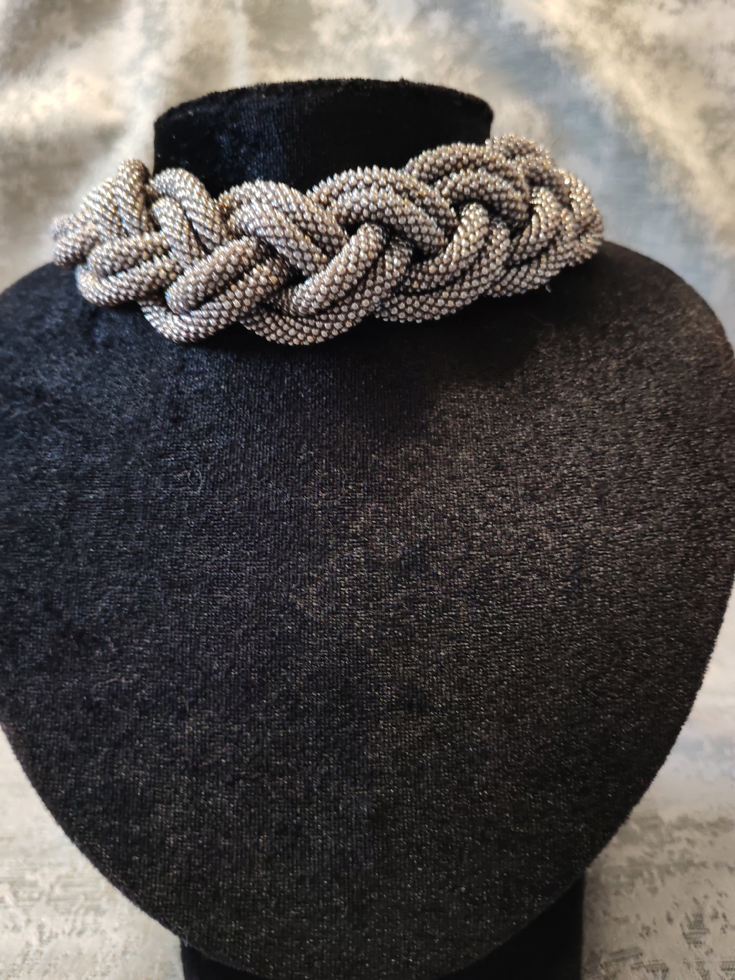 A braided, beaded necklace in shades of silver and gray is displayed around the neck of a black velvet bust form. The necklace is intricately woven, creating a textured, three-dimensional effect. The bust form stands against a muted, patterned background. The lighting is soft, highlighting the details of the necklace. The overall scene is a studio shot focused on showcasing the jewelry.