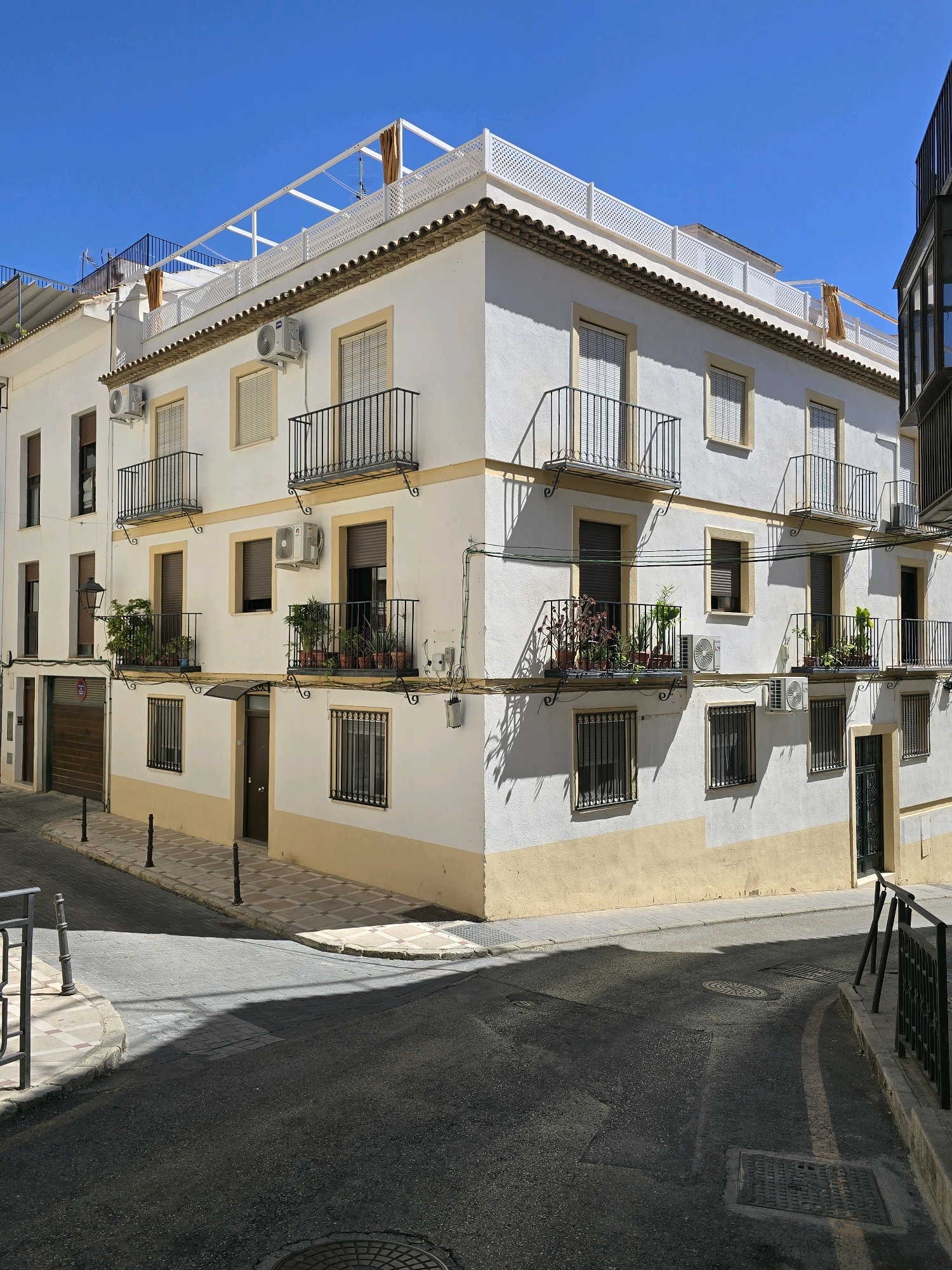 The image captures a sunny day in the city of Jaén, Spain, focusing on a white and yellow multi-story building with traditional balconies. The building is situated on a street with dark asphalt, bordered by a patterned tiled sidewalk. Sunlight casts strong shadows across the street and the building's facade.
The building's architecture features multiple balconies with black wrought-iron railings, adorned with potted plants. Air conditioning units are visible on the exterior walls. The windows are mostly covered with blinds. The ground floor includes a wooden garage door and an entrance with a dark door, framed by barred windows.
The street itself is a key element, showing a curving road with a faint yellow line marking its edge. Pavement covers the sidewalk area, and several black bollards are placed along the edge of the building. A metal fence is visible on the left side of the image. The sky is clear and blue. There are no people or animals visible in the scene.