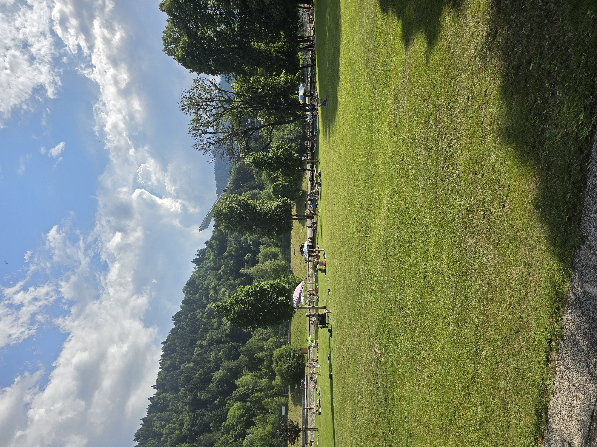 This image depicts the public outdoor swimming pool area in Garmisch-Partenkirchen, Germany, on a sunny day with scattered clouds. The foreground is dominated by a large, well-maintained grassy lawn, with a portion of a gravel path visible in the lower left corner. Numerous individuals are spread across the lawn, engaged in various leisure activities. Some are sunbathing, lying on towels or blankets, while others are sitting or walking. Several colorful umbrellas provide shade for some of the sunbathers. In the middle ground, a shallow pool area is visible, with a few people wading or sitting at the edge. Beyond the pool area, a treeline separates the recreational space from a densely forested hillside that forms the backdrop of the scene. In the far distance, on the left side of the image, a ski jump structure is visible, indicating the proximity to winter sports facilities. The sky is a bright blue, with fluffy white clouds adding texture and depth. The overall atmosphere is one of a relaxed summer day at a popular outdoor leisure venue.