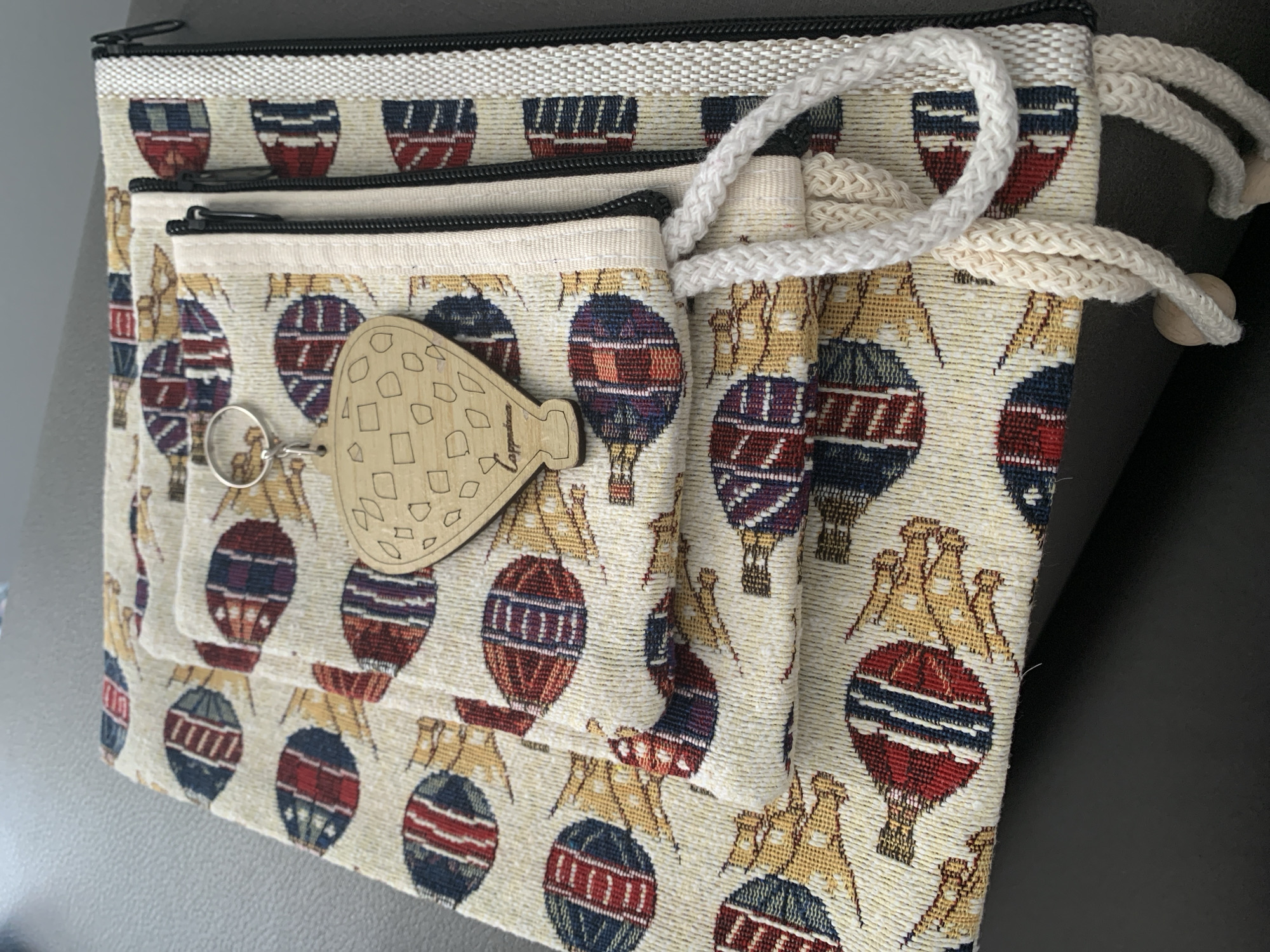The image shows a close-up of a stack of fabric items, possibly pouches or bags, adorned with a repeating pattern of hot air balloons in red, blue, and yellow. A wooden keychain in the shape of a hot air balloon with cutouts and the word "Cappadocia" is attached to one of the items. A white braided rope handle is also visible. The items are placed on a dark, textured surface, likely a table. The background is blurred, but appears to be indoors. There is no indication of the city of Antalya, Turkey, in the image.