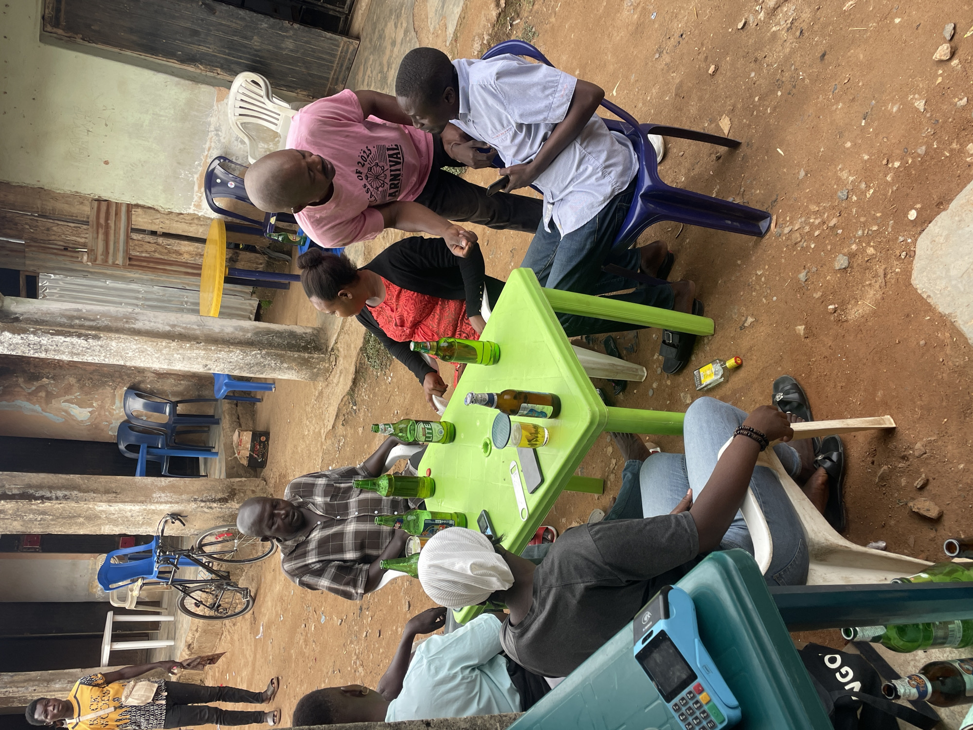 A group of people are gathered around a light green plastic table outdoors in Jos, Nigeria. Six individuals are partially visible, seated in plastic chairs of various colors (blue, white, light green). The table is covered with multiple green beer bottles, a glass, a bottle opener, and a mobile phone. A woman in a red and black top is sitting at the table. Two men are standing and looking at a mobile phone held by one of them. One is wearing a pink t-shirt. A bicycle is visible in the background against a wall. Another man, in a brown plaid shirt, sits at the table. A point-of-sale (POS) machine is placed near the table edge. Several discarded beer bottles lie scattered on the ground. The environment suggests a casual outdoor setting, possibly a local bar or gathering place. The ground is dirt.