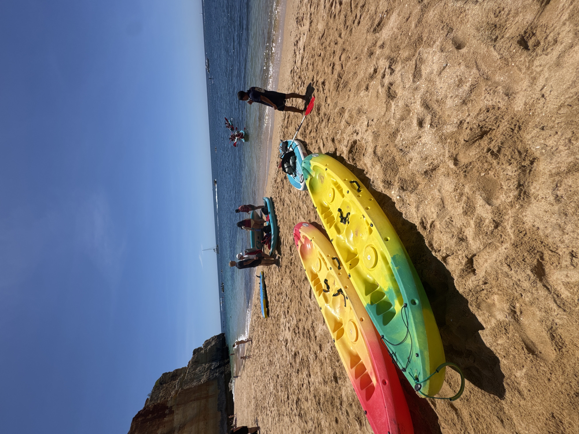 The image shows a beach scene. Two kayaks, one yellow and one red, lie on the sand in the foreground. In the midground, there are several people near the water's edge, some standing and some on paddleboards or kayaks. A person stands near the water with a paddle. Further out, there are sailboats on the water. The sky is blue and clear. A cliff is visible on the left side of the frame. The location is the city of Unknown, Unknown.