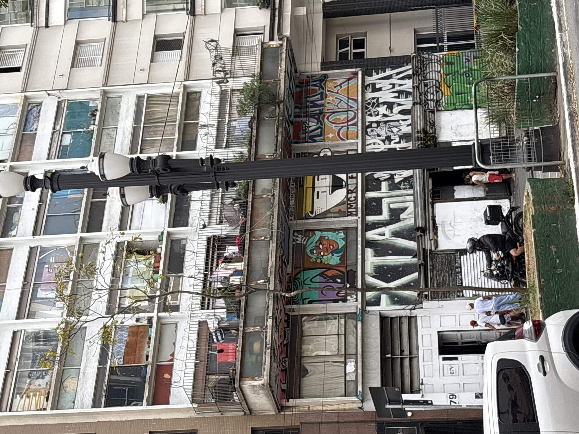 The image depicts a street scene in São Paulo, Brazil, likely in a dense urban area. A multi-story building dominates the frame, characterized by its utilitarian architecture, numerous windows, and balconies adorned with laundry. The lower facade of the building is heavily decorated with vibrant graffiti, showcasing various artistic styles.
In the foreground, a white vehicle is partially visible on the left. To the right of the vehicle, three individuals are walking near an entrance labeled "79" and "879". One individual is wearing a red cap and shorts, another is in a white t-shirt and light blue pants, and the third is partially obscured. A black motorcycle is parked nearby, with a person wearing a helmet and dark clothing seated on it.
A black lamppost with multiple fixtures stands prominently in the center of the image, partially obscuring the building's facade. Lush green vegetation is visible in planters along the street. The overall impression is one of a busy, lived-in urban environment, marked by both decay and vibrant artistic expression.